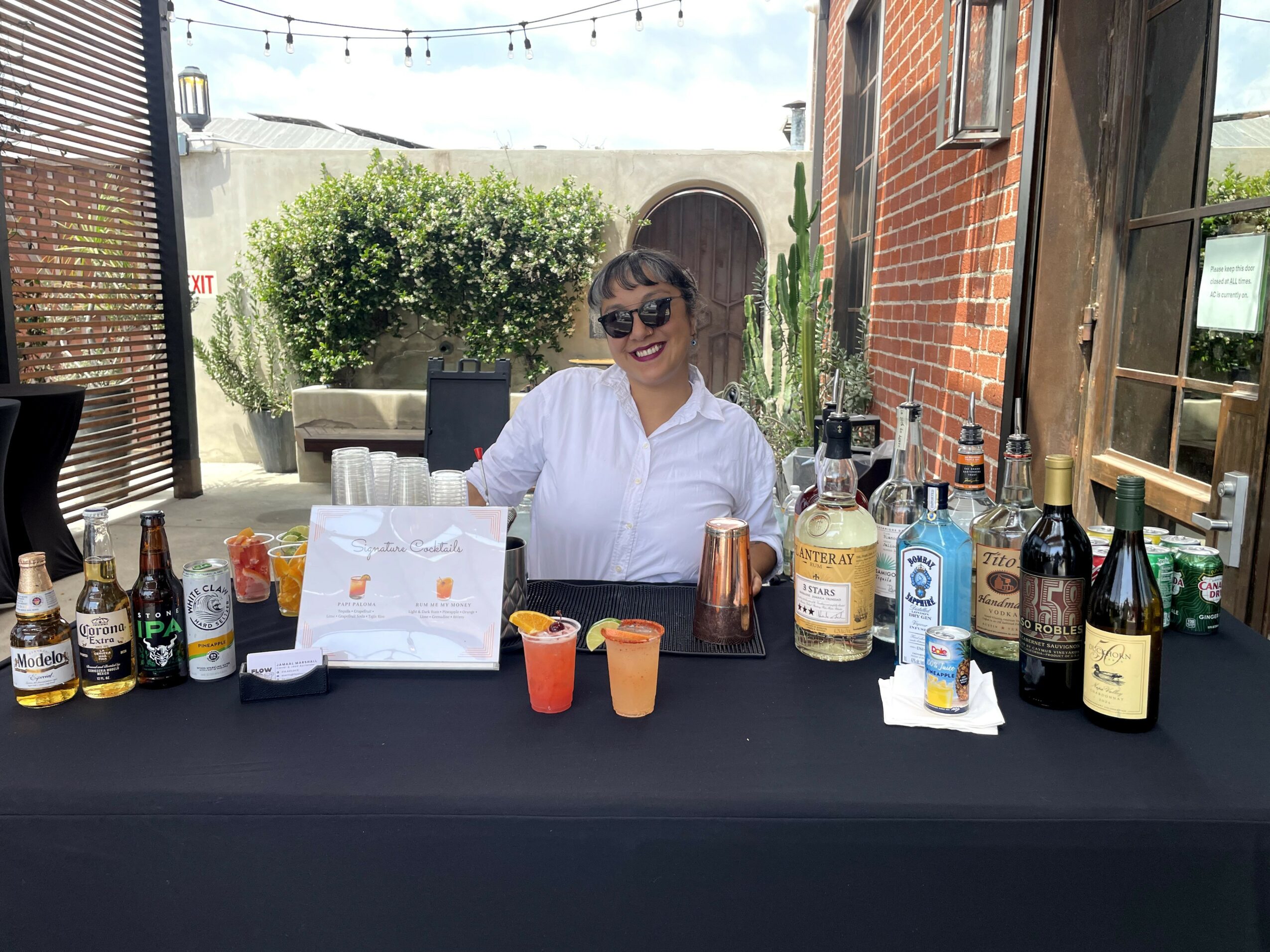 Woman with sunglasses behind a bar table with various bottles and drinks outdoors, brick wall and greenery in background.
