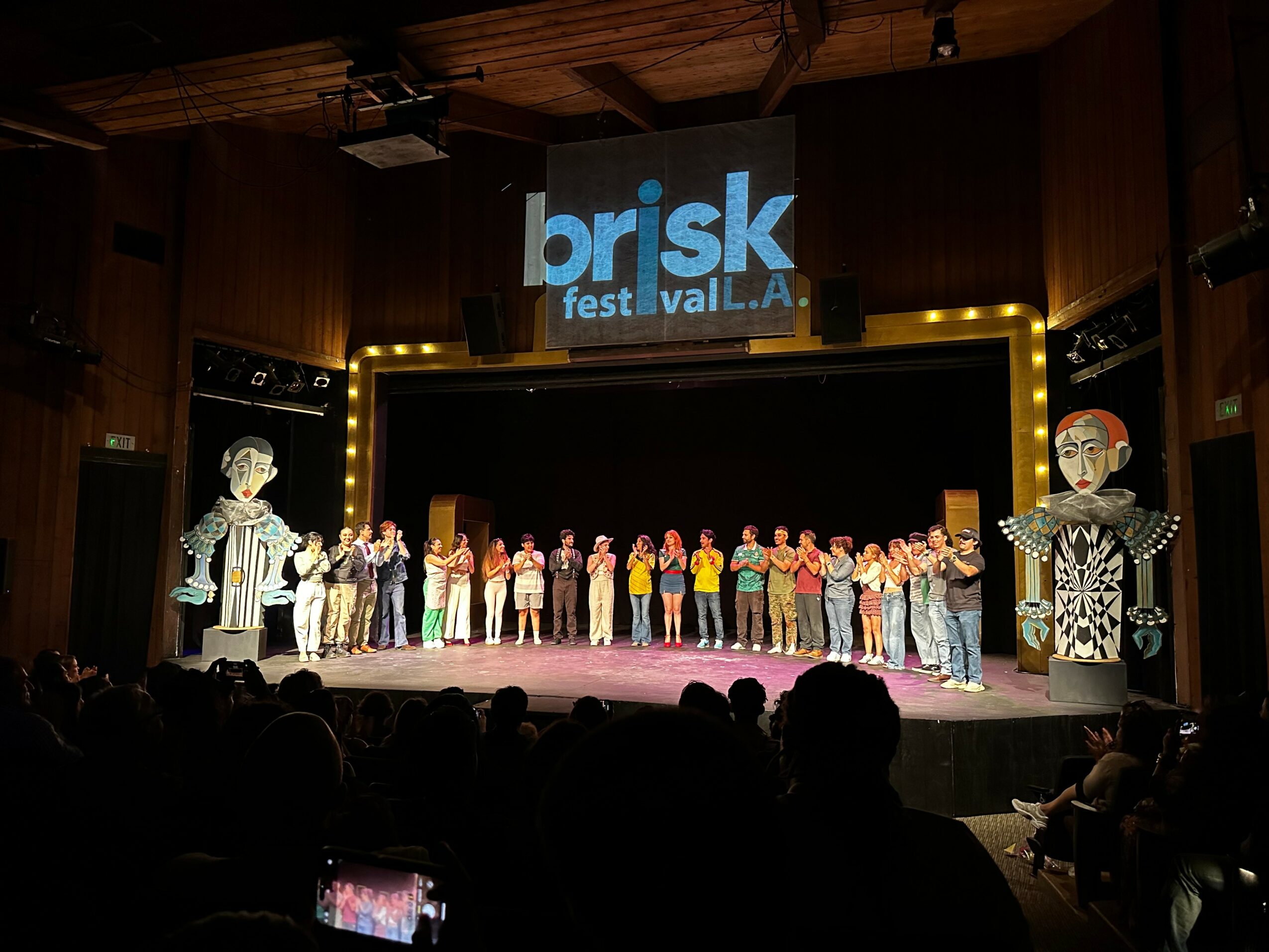 Group of performers on stage with large decorative figures, stage framed by lights, audience in foreground, festival banner above.