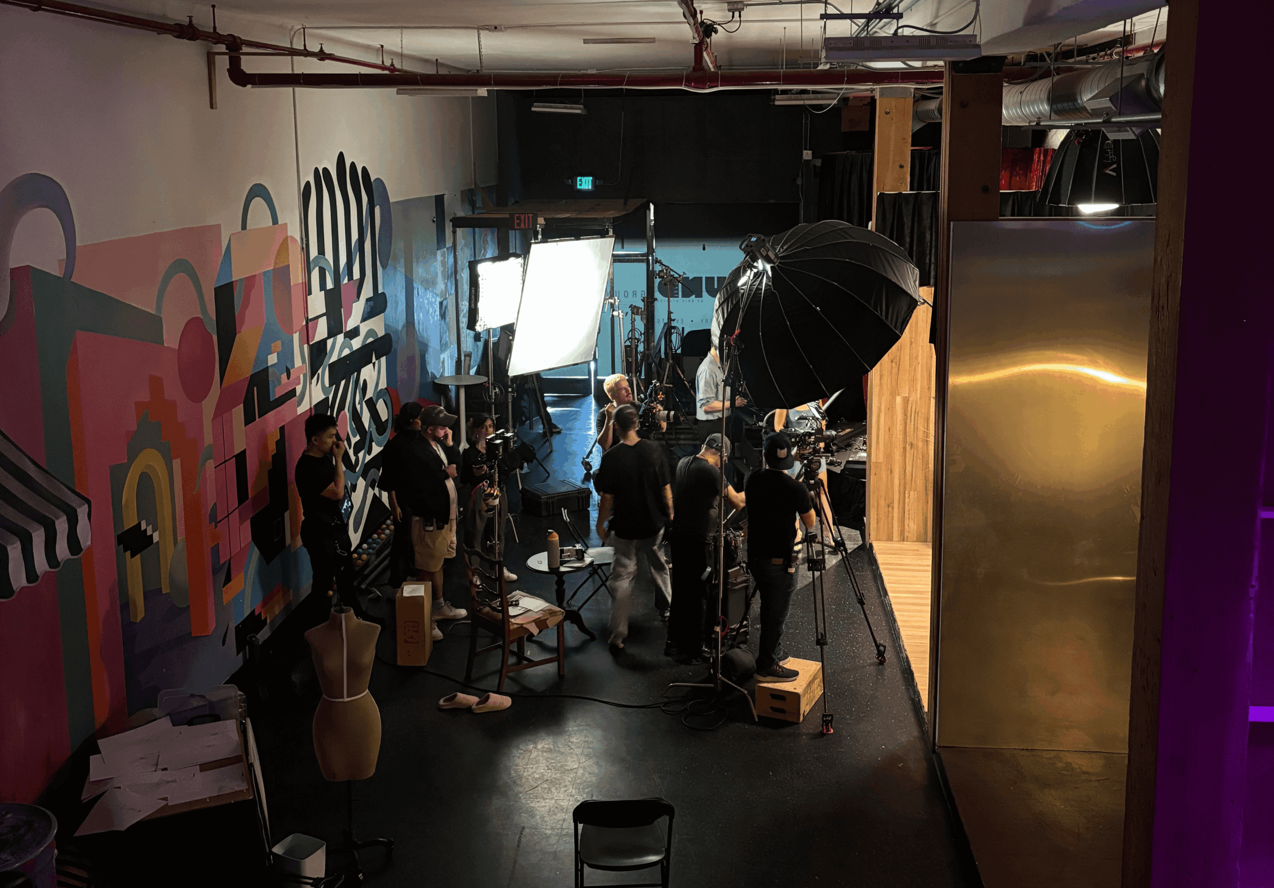 People gather in a studio with colorful wall art, lighting equipment, and a large black umbrella, preparing for a shoot.