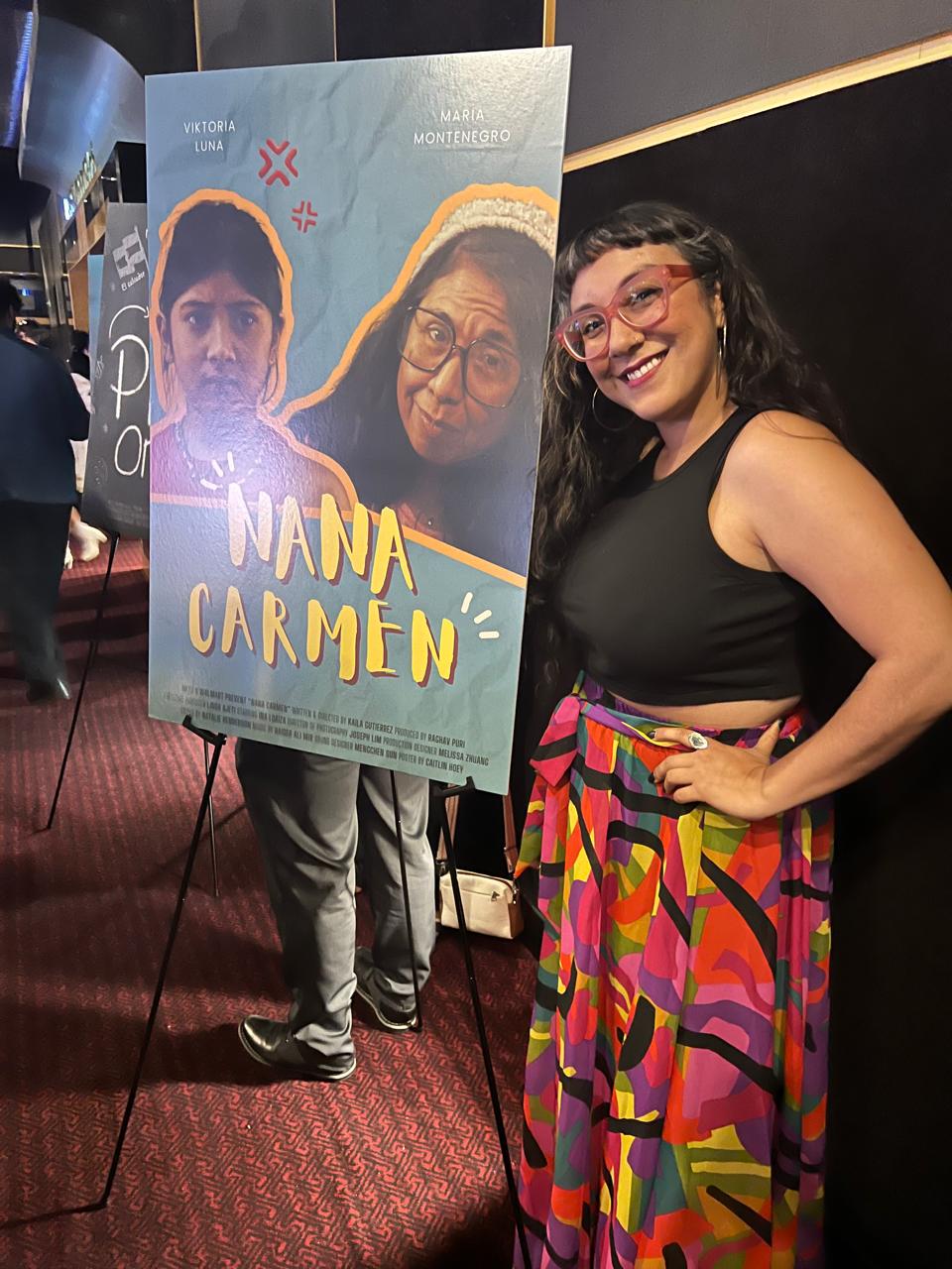 Woman smiling in black top and colorful skirt standing next to a poster for 'Nana Carmen' with two faces on it.