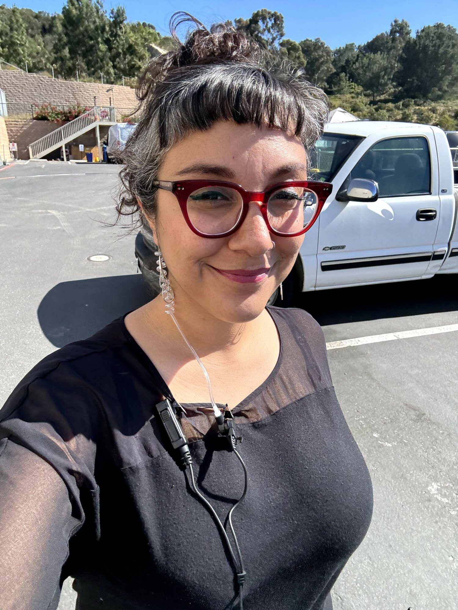 Woman with glasses and earrings smiling outdoors in a parking lot, with trees and a building in background.