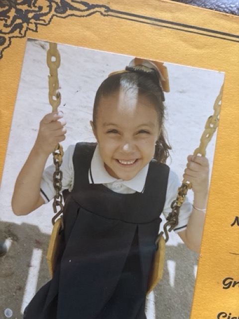 Young girl smiling on a swing, holding the chains, outdoors with a light background.