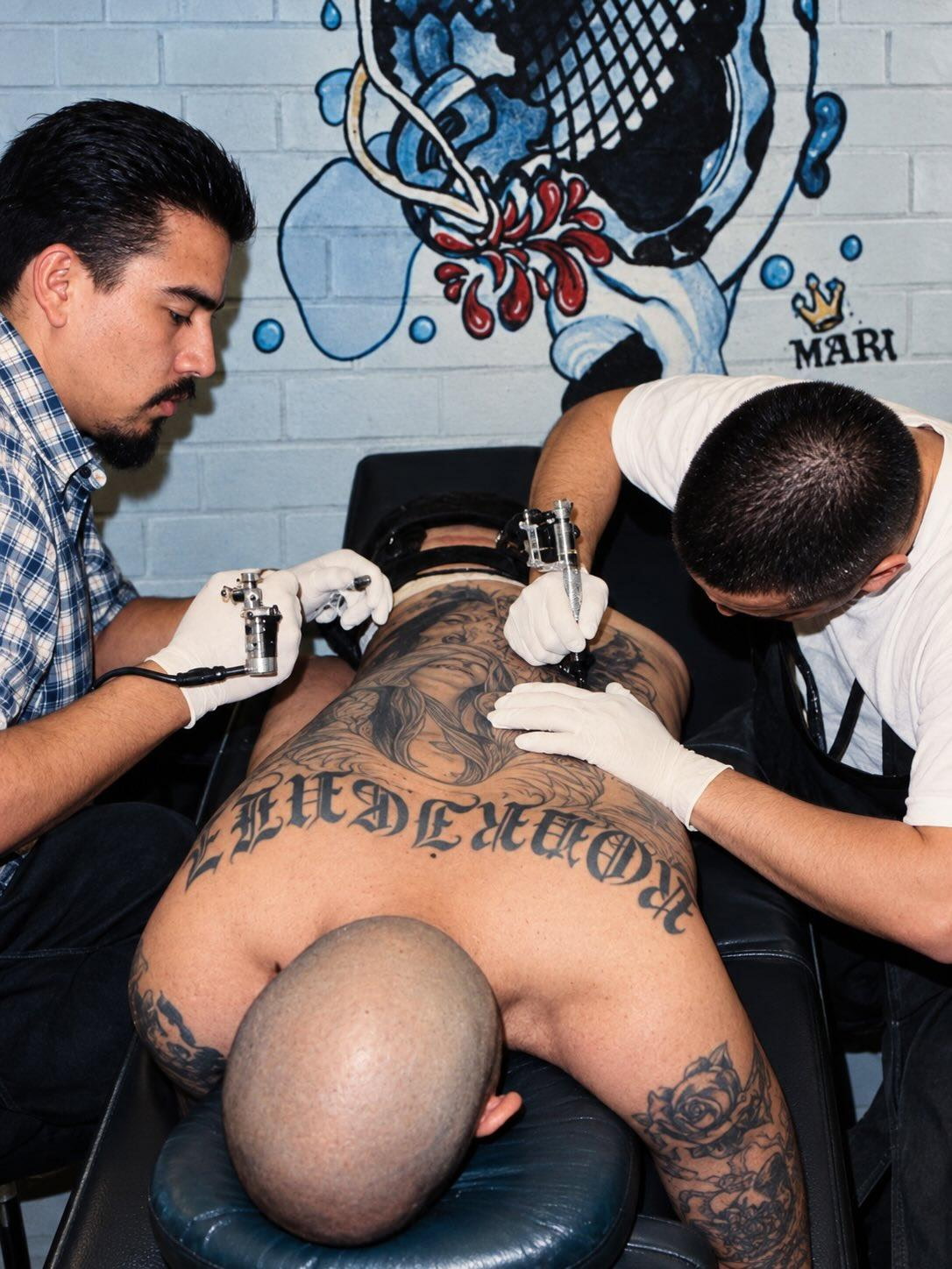 Two tattoo artists work on a person's back in a studio with a mural background.