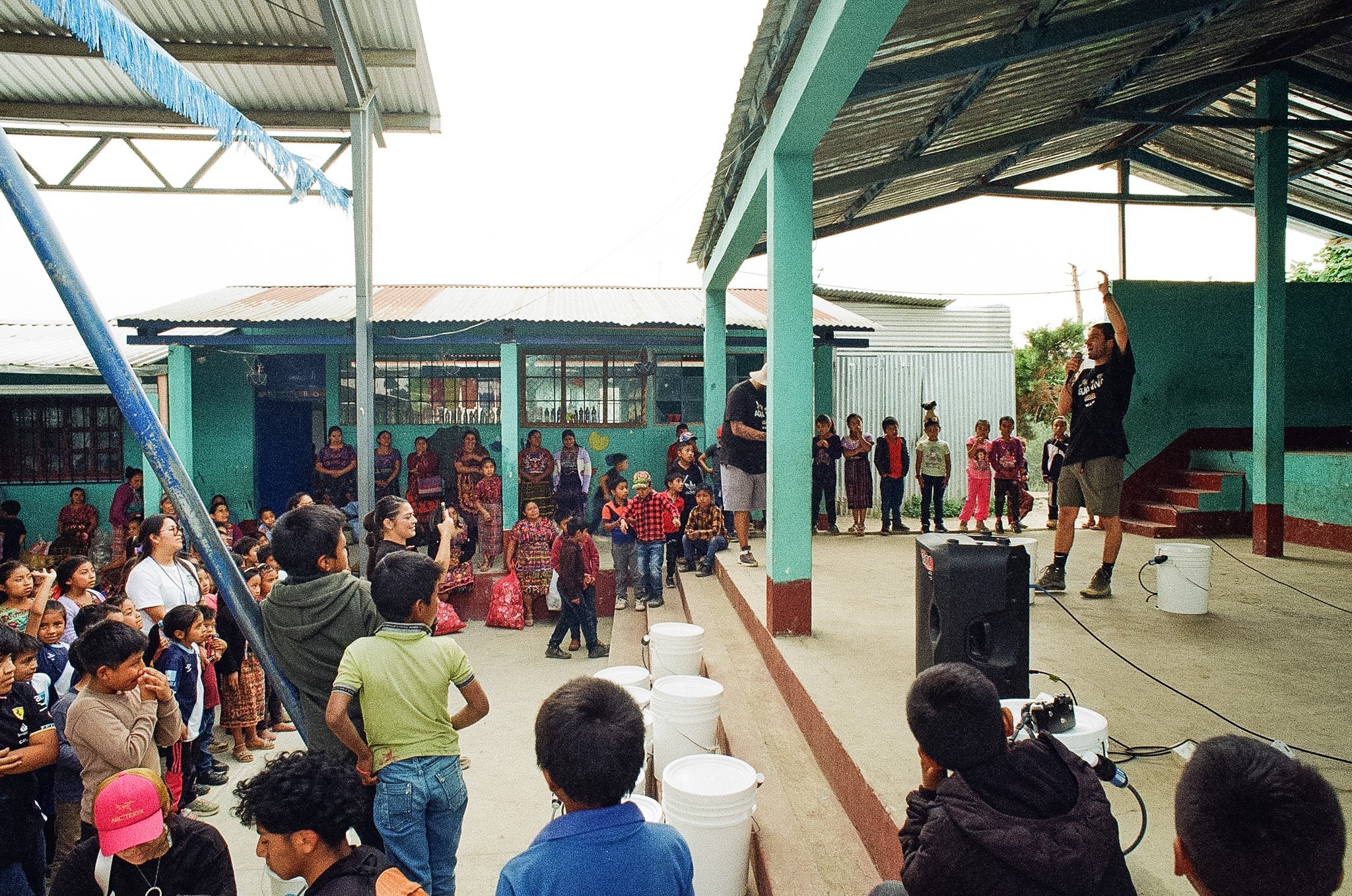 Children and adults gather outdoors under a covered area, some sitting and some standing, with a speaker on stage.