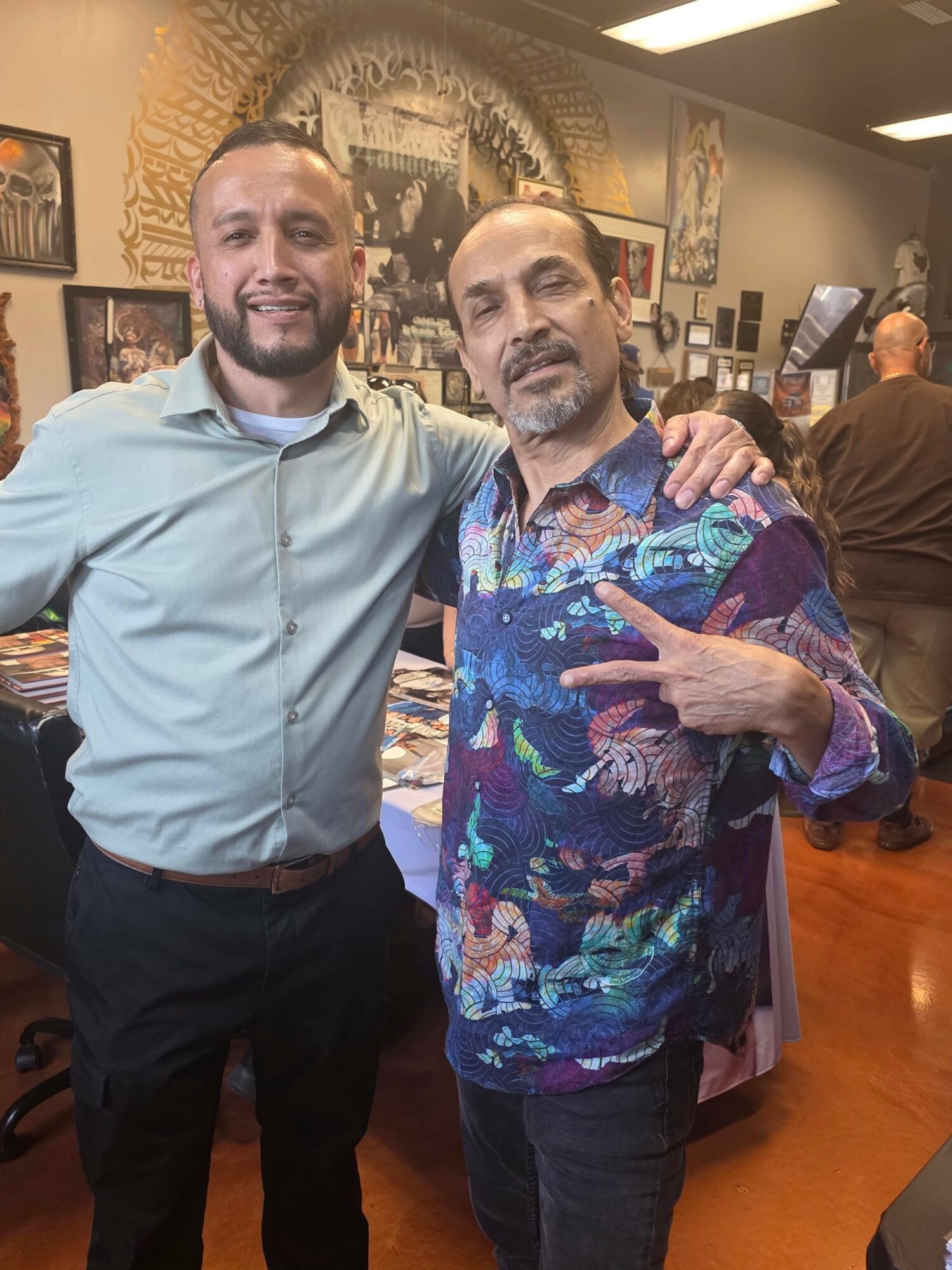 Two men standing close together, smiling, one with arm around the other, in an art gallery or store with artwork on walls.