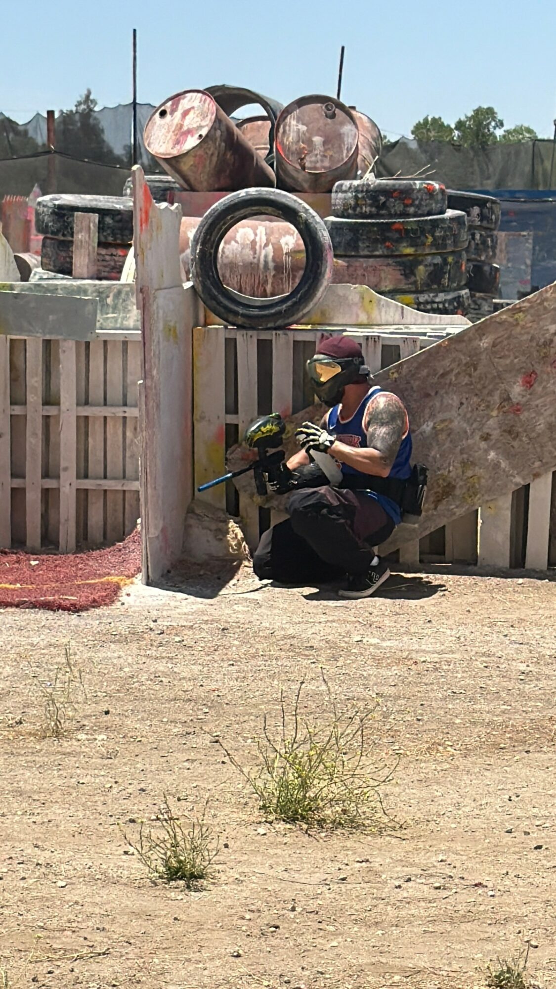 Person crouching behind a wooden barrier, holding a paintball gun, wearing protective gear, in an outdoor paintball field.