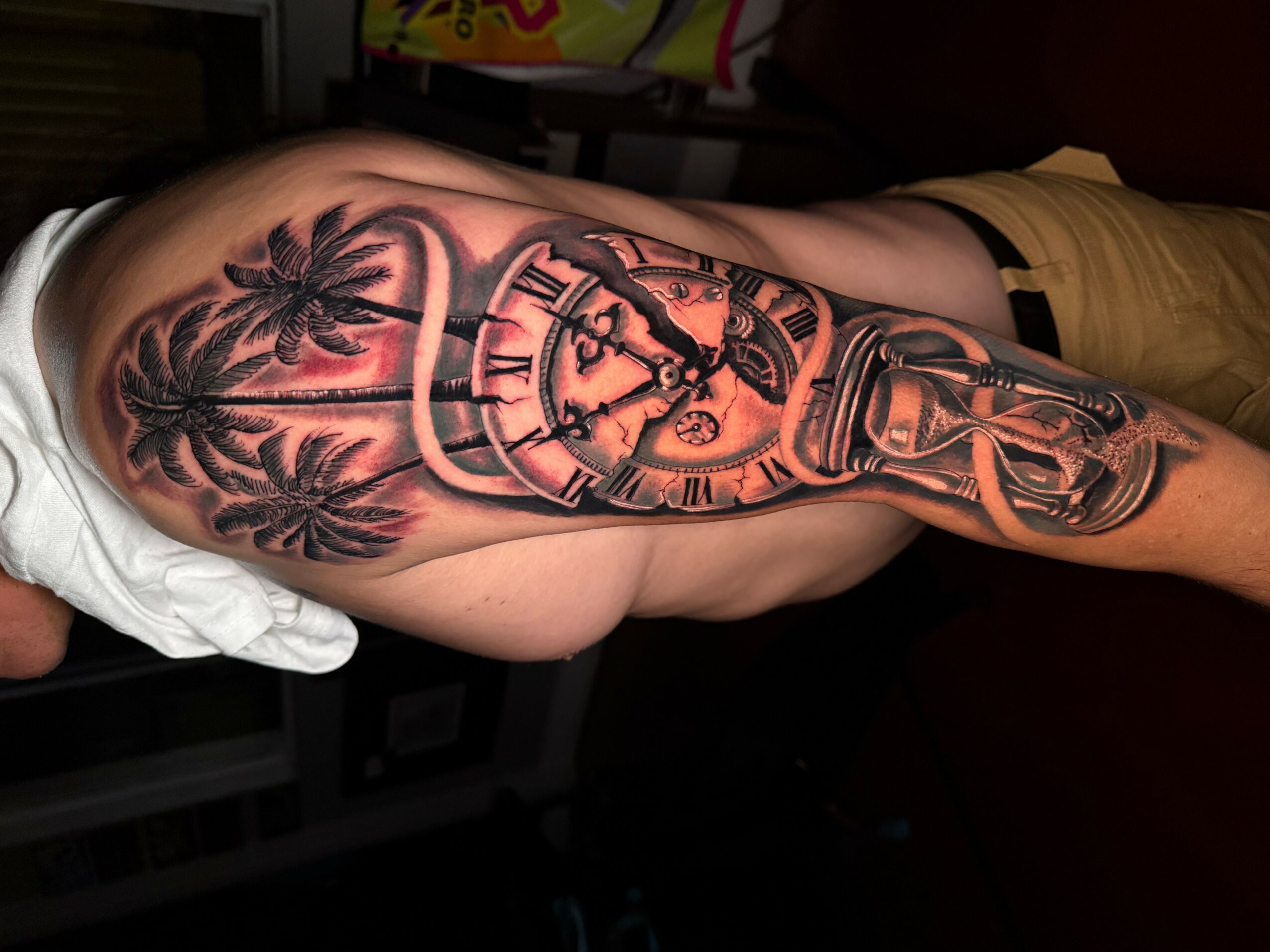 Tattoo of a pocket watch with palm trees on upper arm, detailed and shaded, with a background of dark skin.