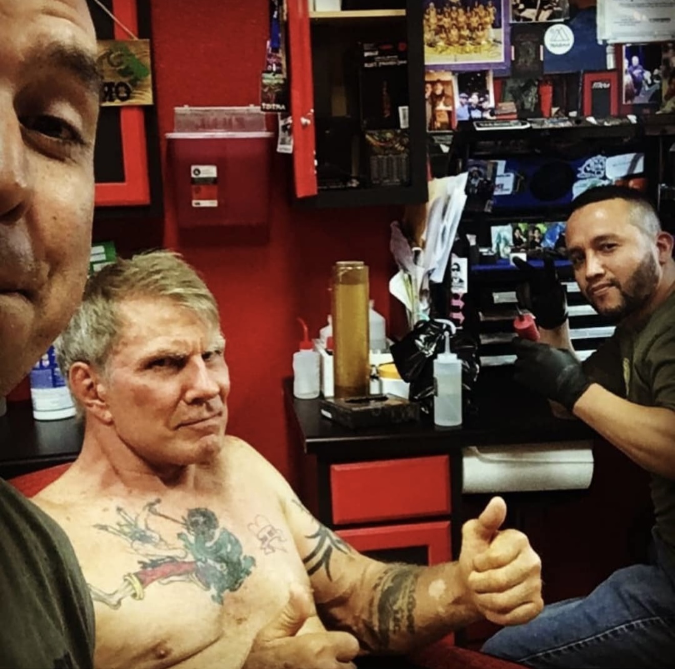 Three men in a tattoo shop, one shirtless with tattoos, giving a thumbs-up, and two others working behind the counter.