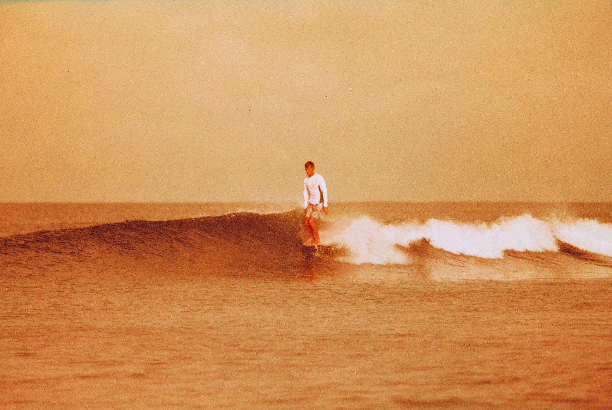 Person standing on a small wave in the ocean, with a yellowish sky in the background.