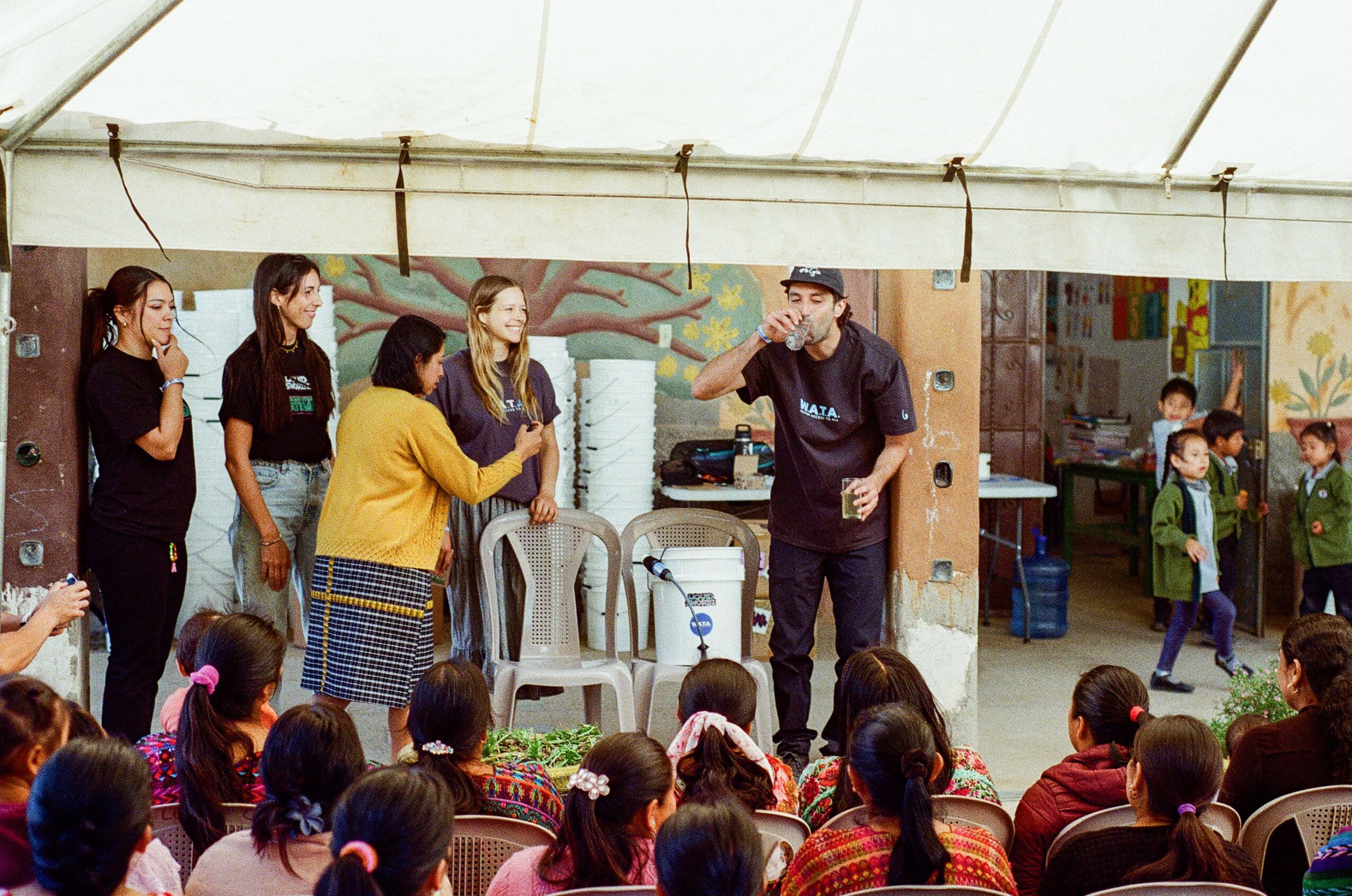 A man demonstrates a skill to a group of children and adults under a tent, with children seated and watching attentively.