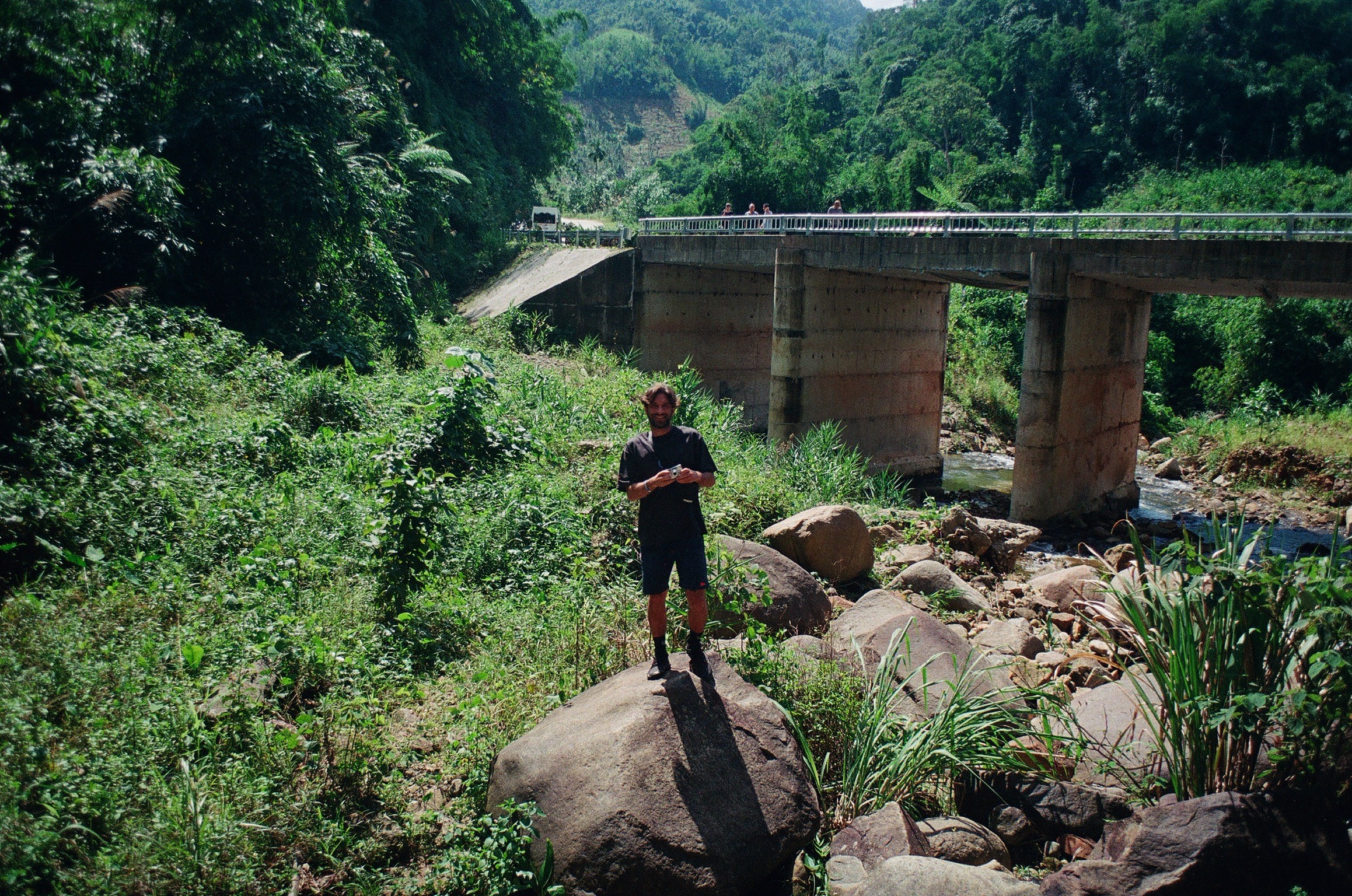 Person standing on a large rock near a river with a bridge overhead, surrounded by greenery and trees.