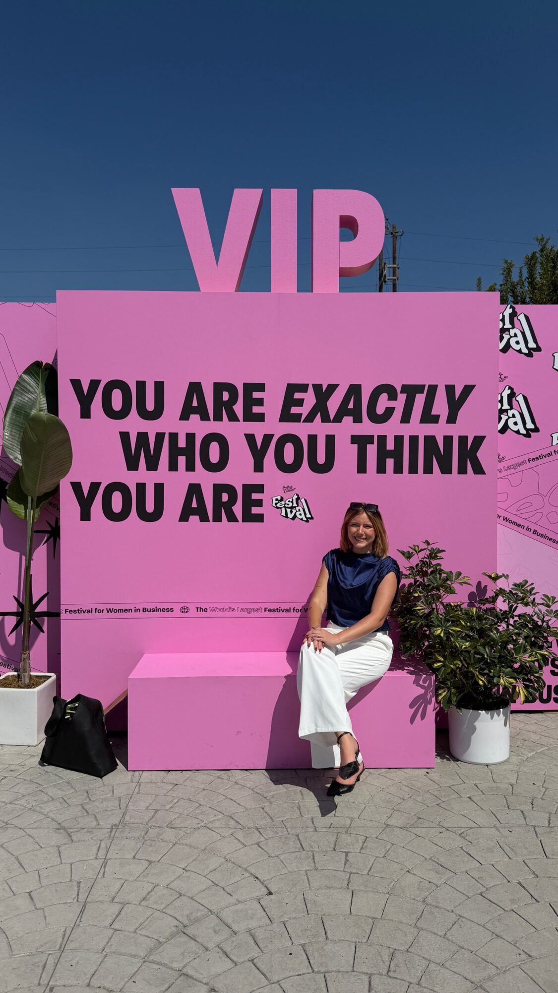 Woman sitting on pink bench in front of pink wall with large VIP sign and motivational quote, outdoors.