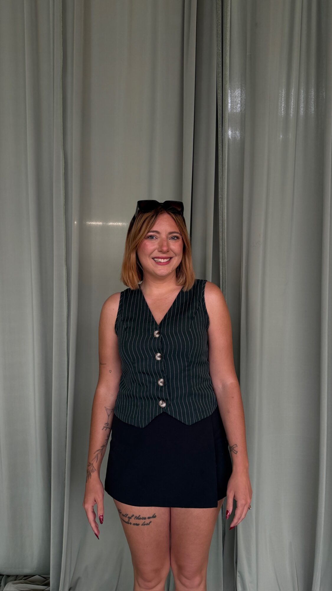 Woman with shoulder-length hair smiling, wearing a sleeveless vest and dark skirt, standing in front of curtains.