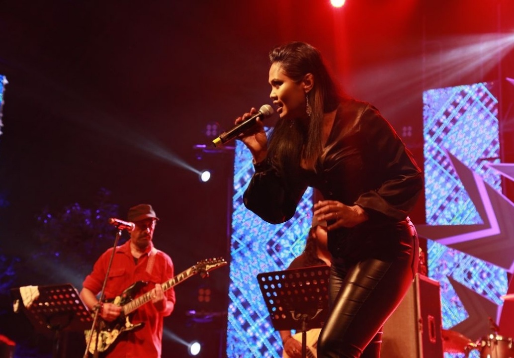 Female singer performing with a male guitarist on stage, colorful background with lights and patterns, stage setting, music performance