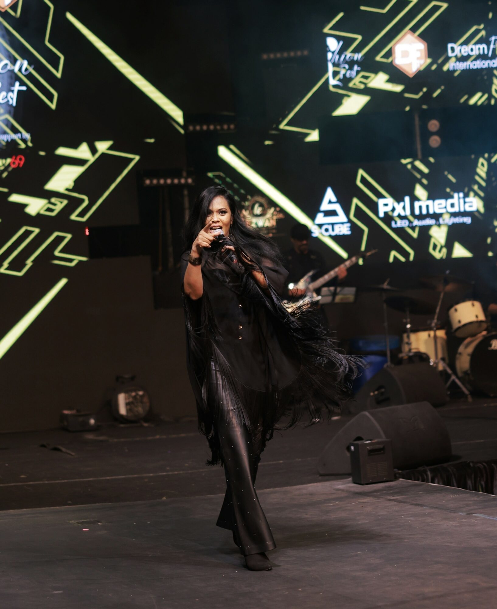 Performer singing on stage with digital graphics in background, holding a microphone, wearing dark clothing, and dancing.