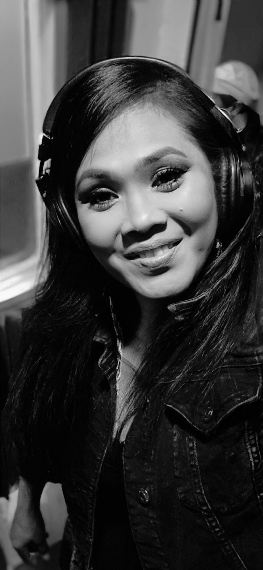 Young woman with long dark hair wearing headphones and a denim jacket, smiling at the camera.