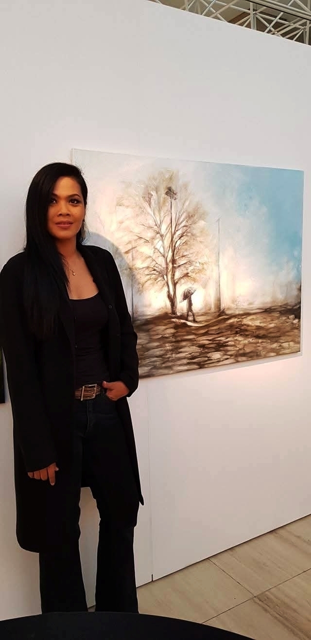 Woman standing next to a landscape painting of a tree and a person, in an indoor gallery setting.