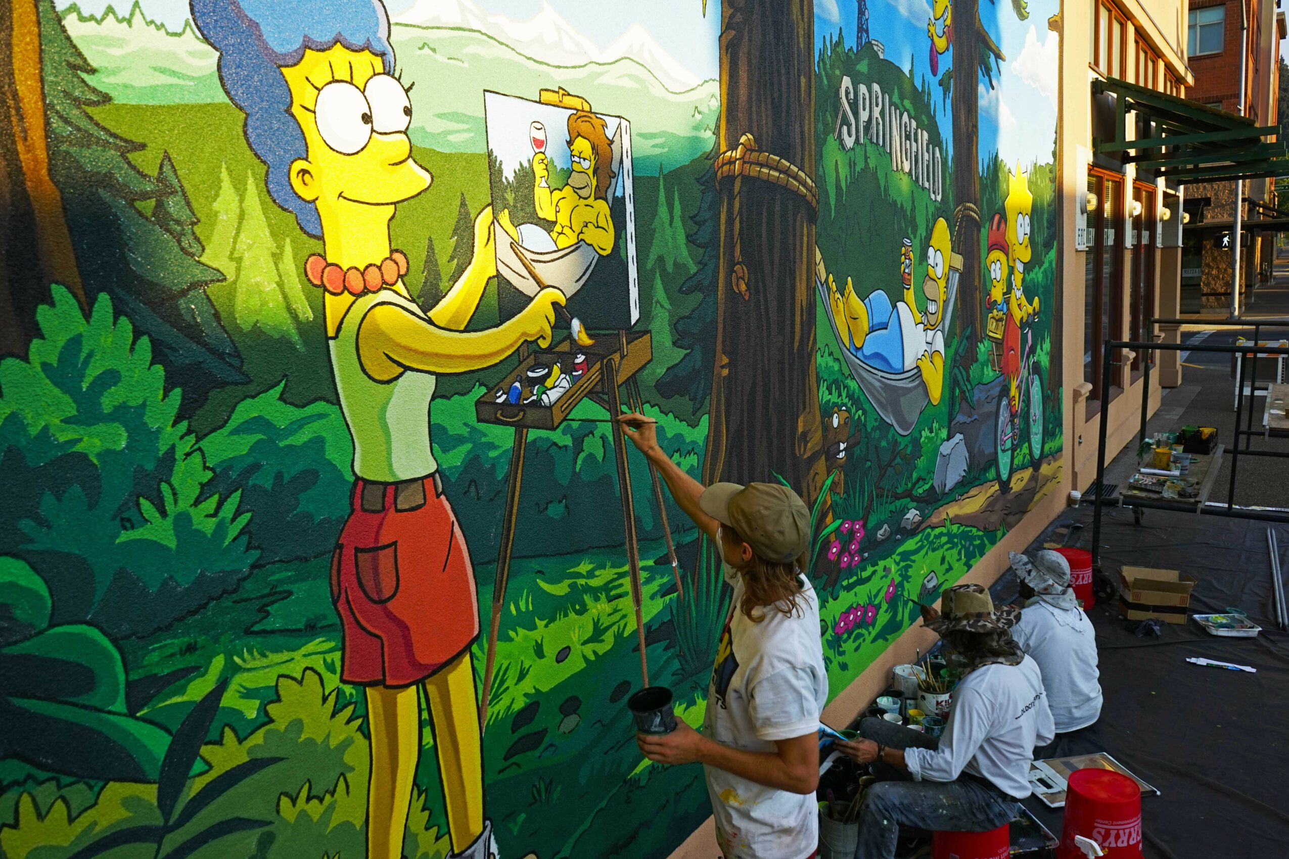 People painting a mural of Marge Simpson from The Simpsons on a colorful outdoor wall.