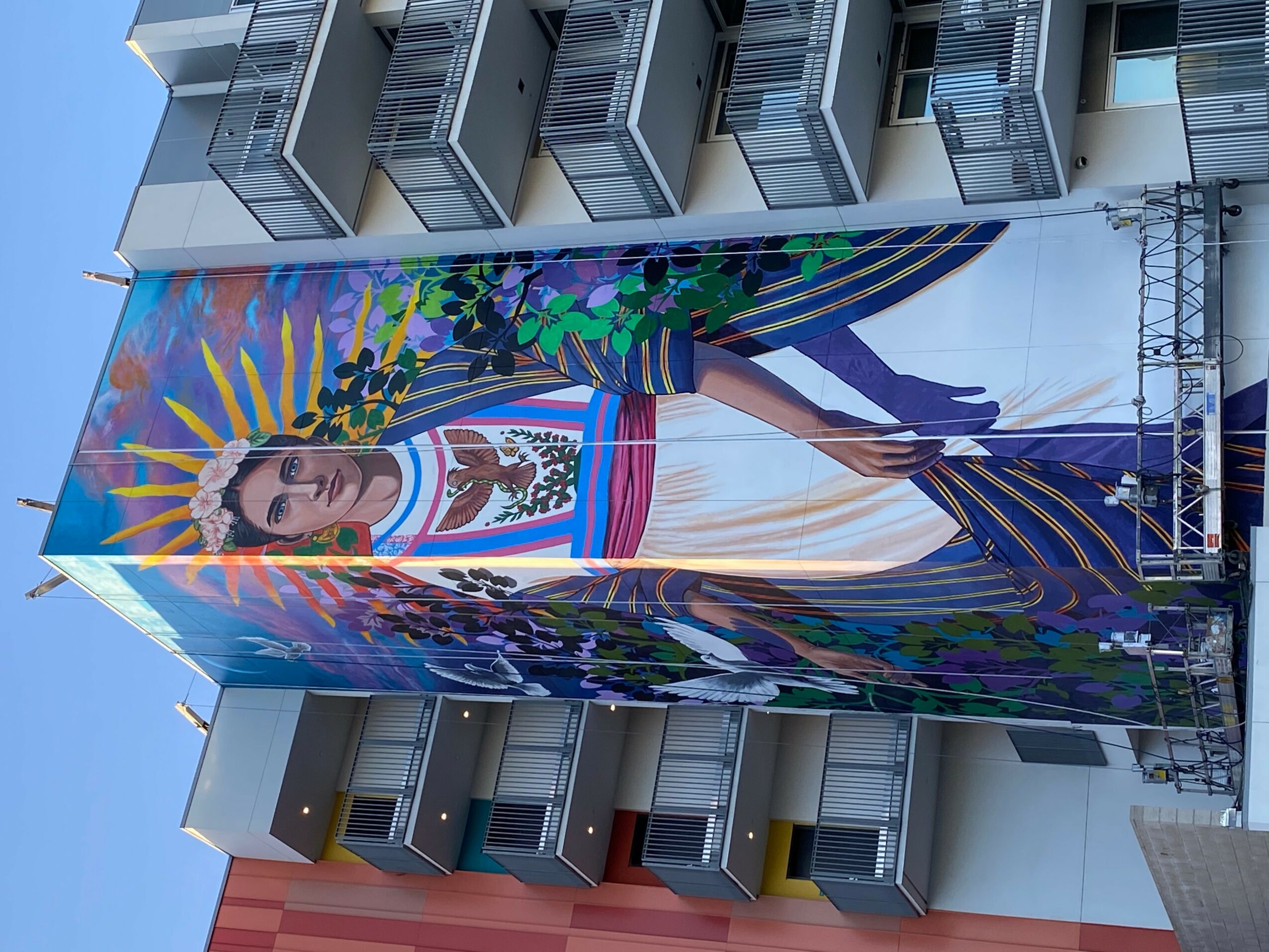 Large colorful mural of a woman with a headdress, surrounded by vibrant patterns, on the side of a building.
