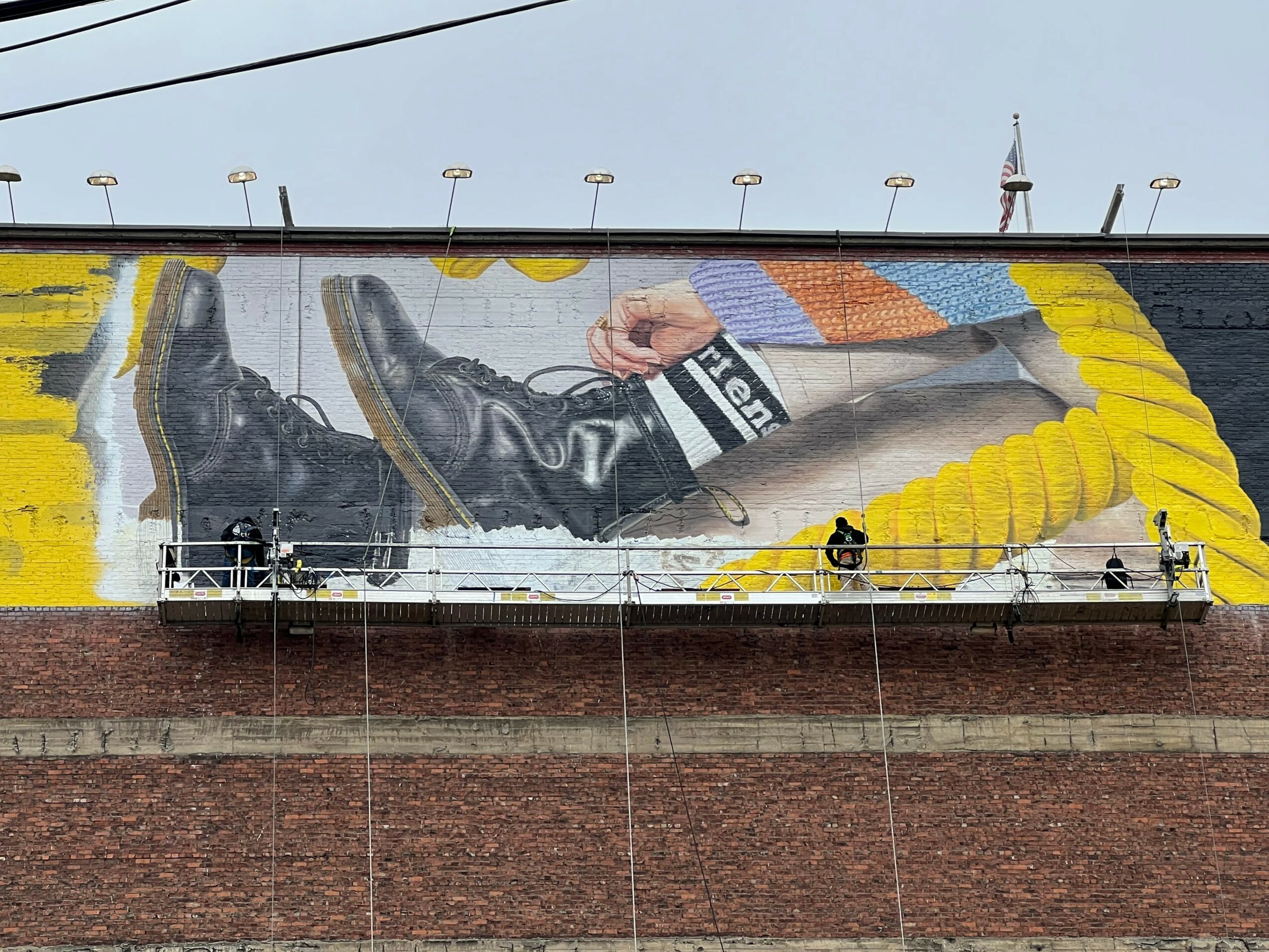 Large mural of a person lying down, wearing black shoes and pants, with yellow and orange elements, on a brick wall.