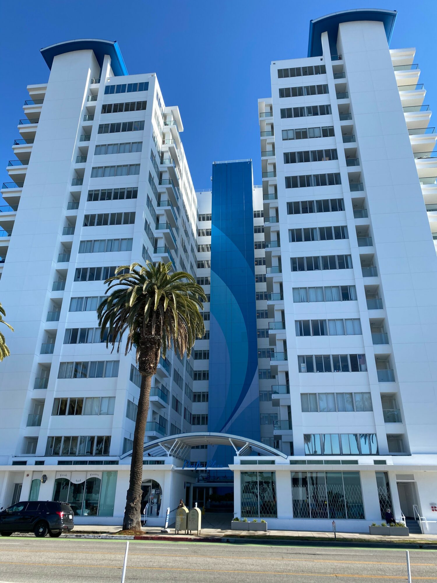 Two tall white modern buildings with blue accents and a curved blue sculpture in front, under a clear blue sky.