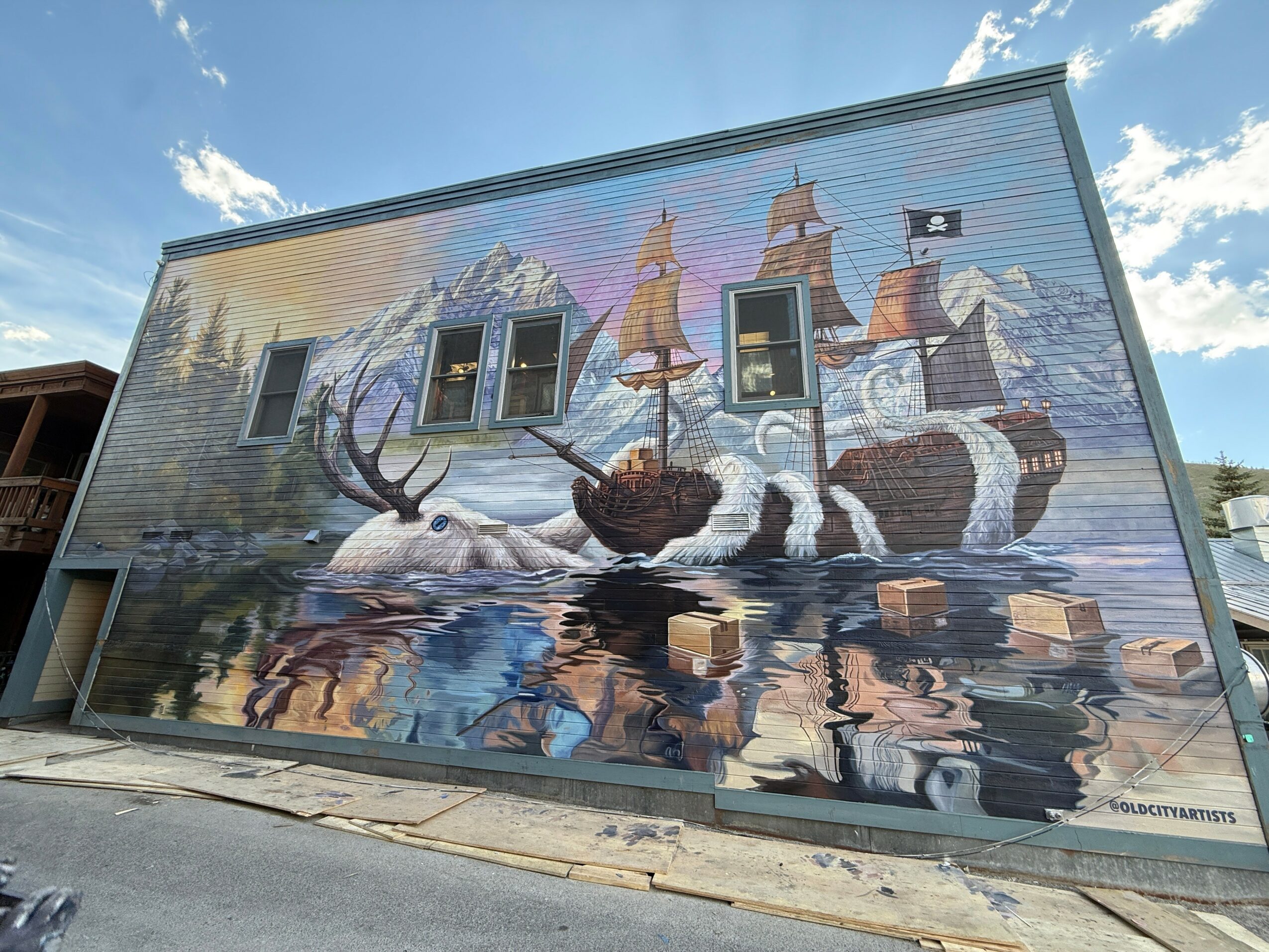 Large mural of a sailing ship with snowy mountains and water reflections on building wall, under a partly cloudy sky.