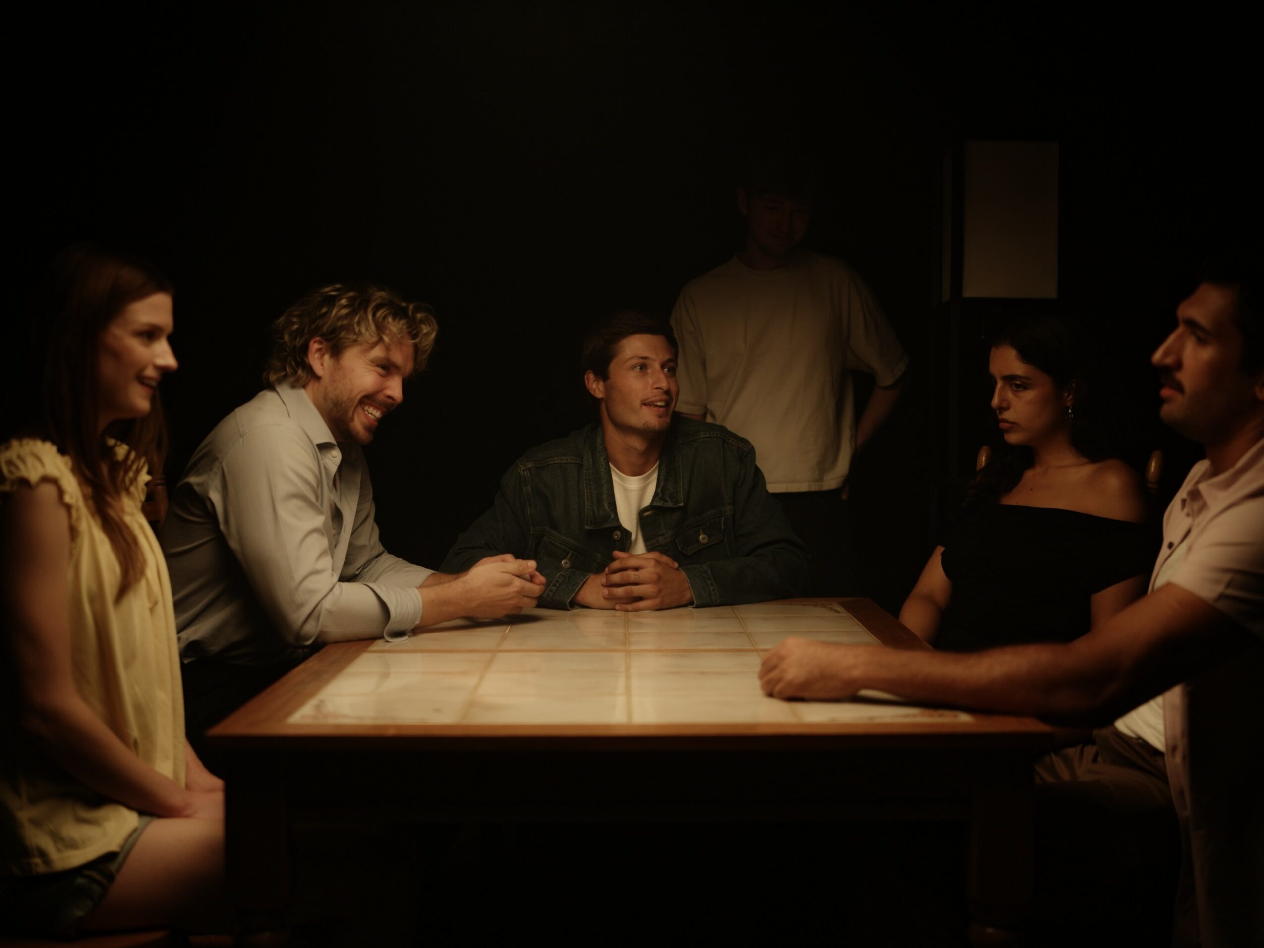 Six people sit around a table in a dark room, engaging in conversation, with one person standing in the background.