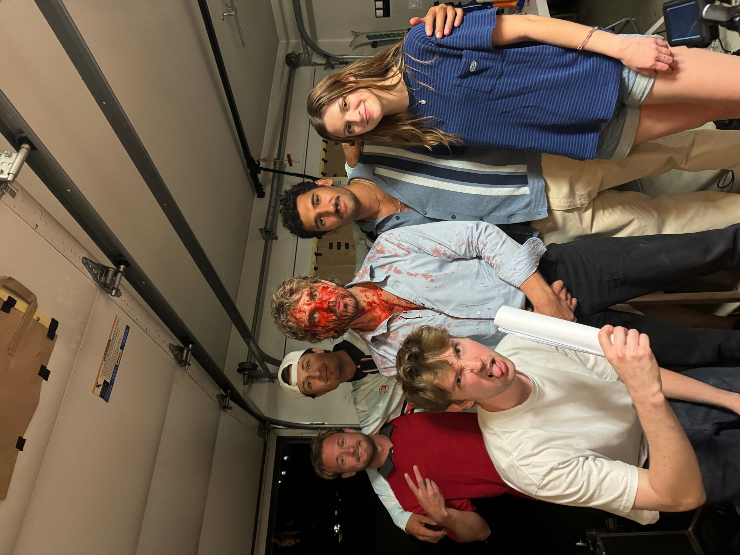 Group of six people posing indoors, some making peace signs, with a ceiling and equipment visible in the background.