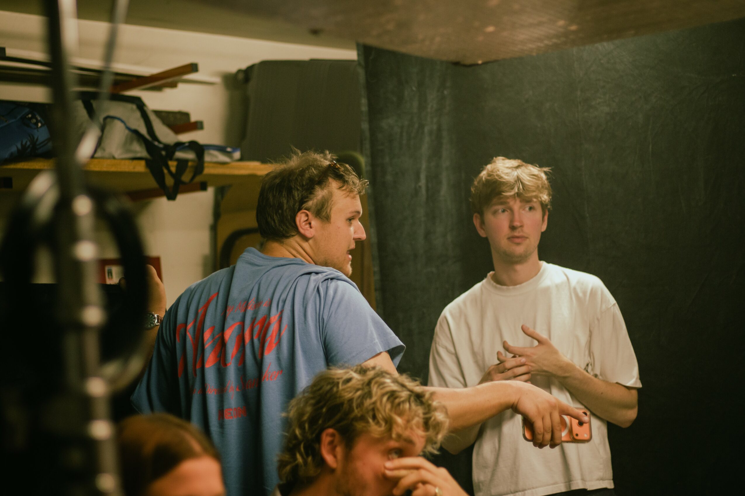 Two young men are in a room with a dark backdrop, one with a blue shirt and the other with a white shirt, engaging in conversation.