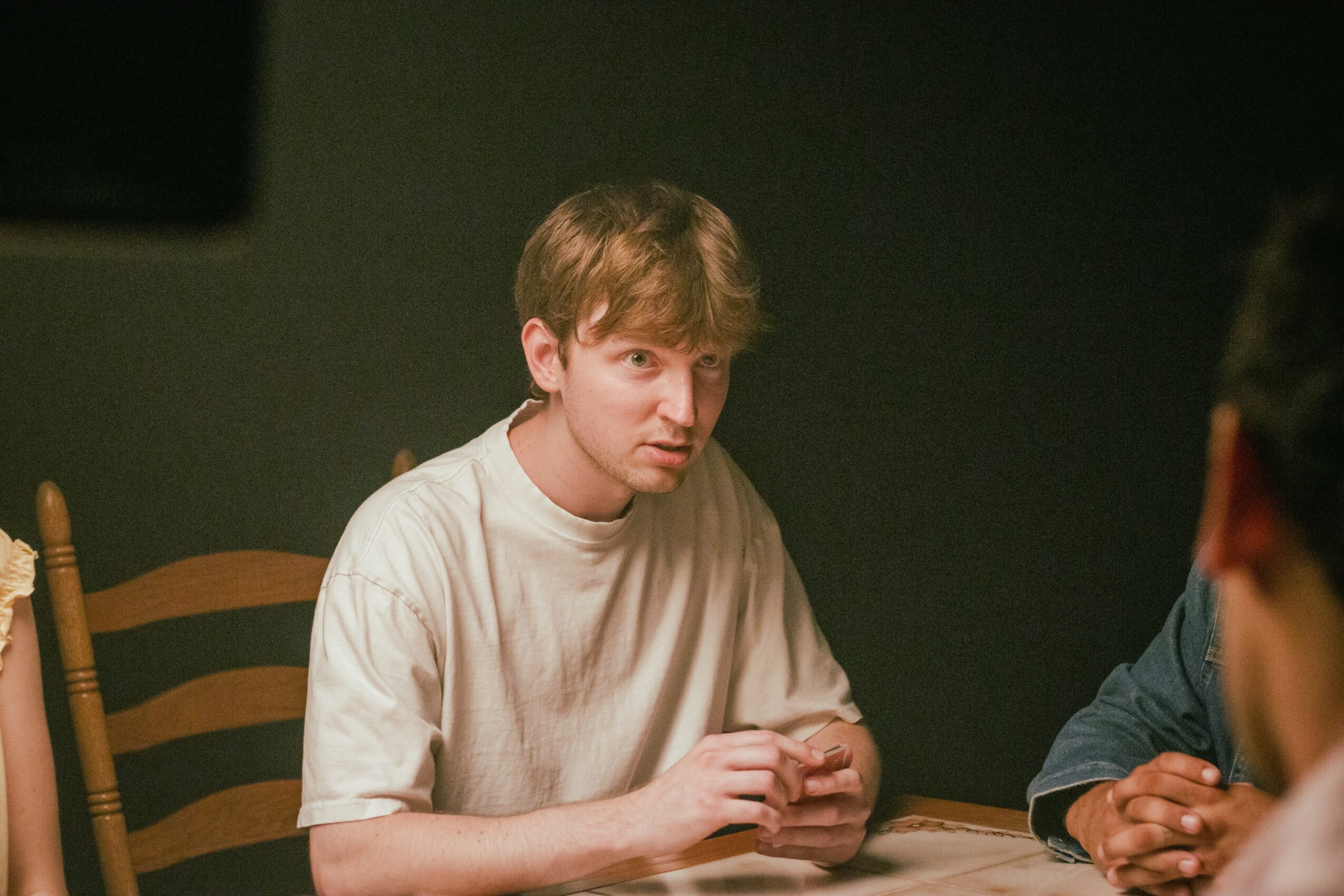 Young man with light brown hair sitting at a table, wearing a white shirt, with a dark background.