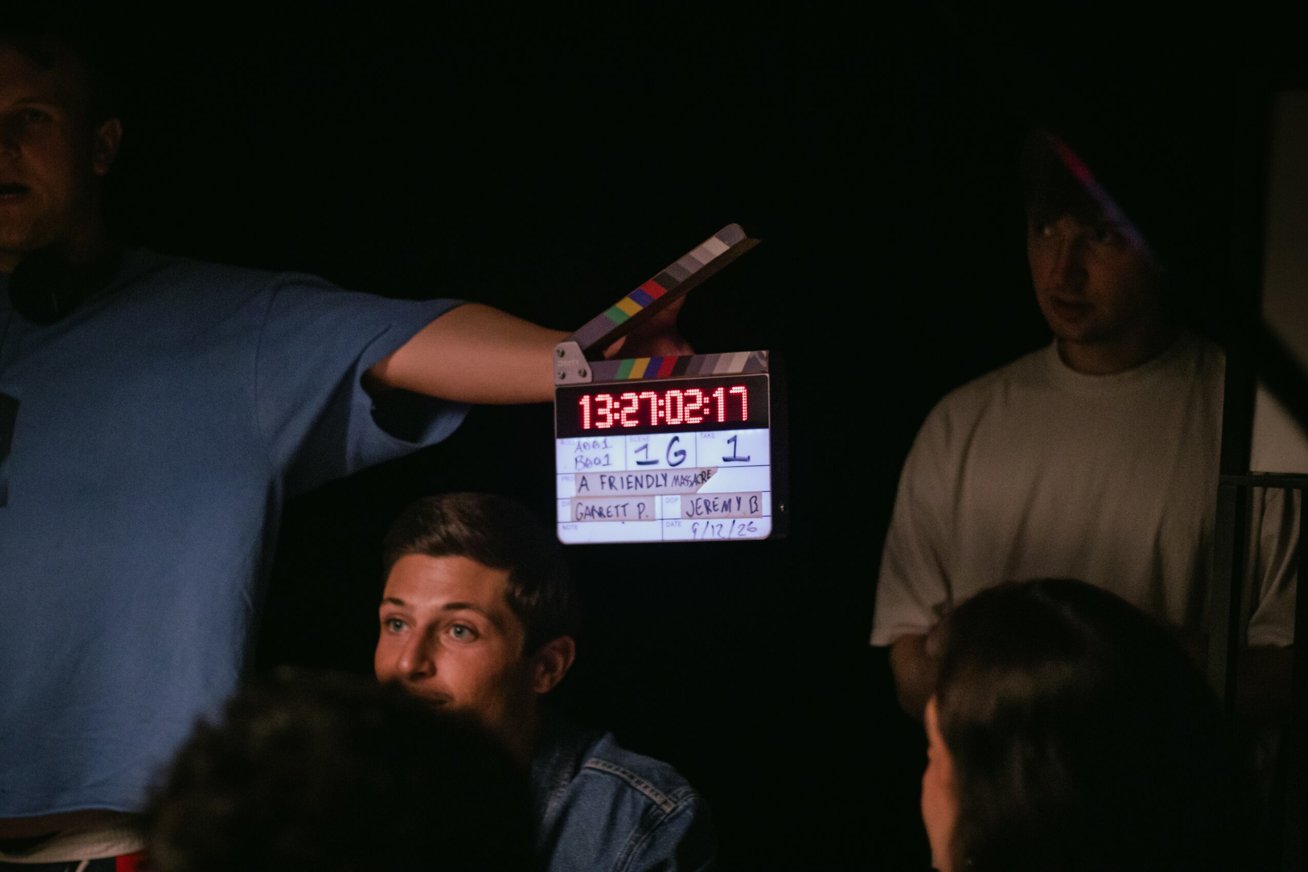 People in a dark room, one person holding a clapperboard, others seated and standing, some faces visible, some obscured.