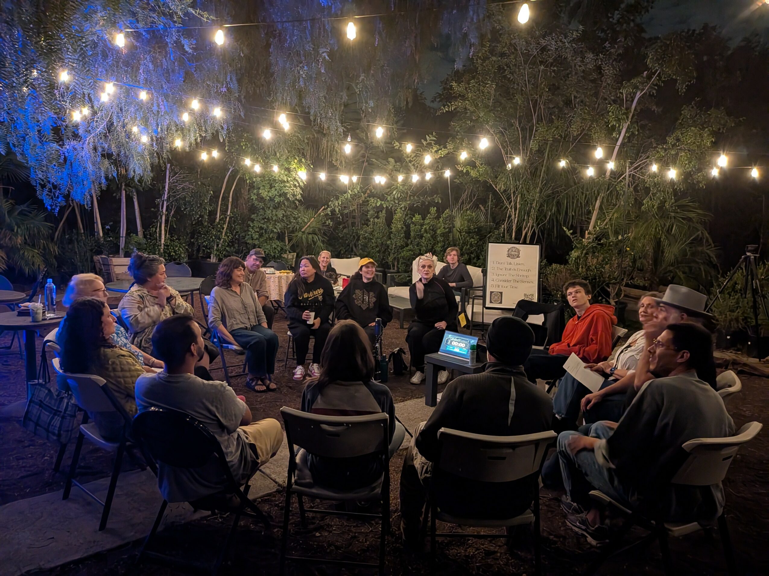 Group of people sitting outdoors under string lights at night, engaged in a discussion or presentation.