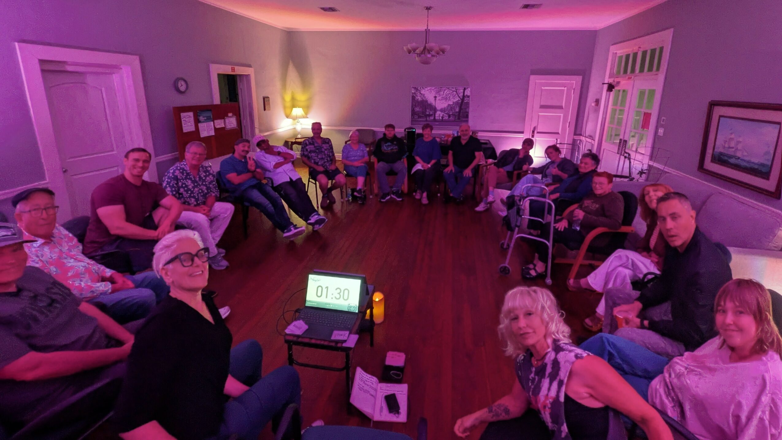 Group of people seated in a circle in a room with pink lighting, some facing the camera, some turned away.