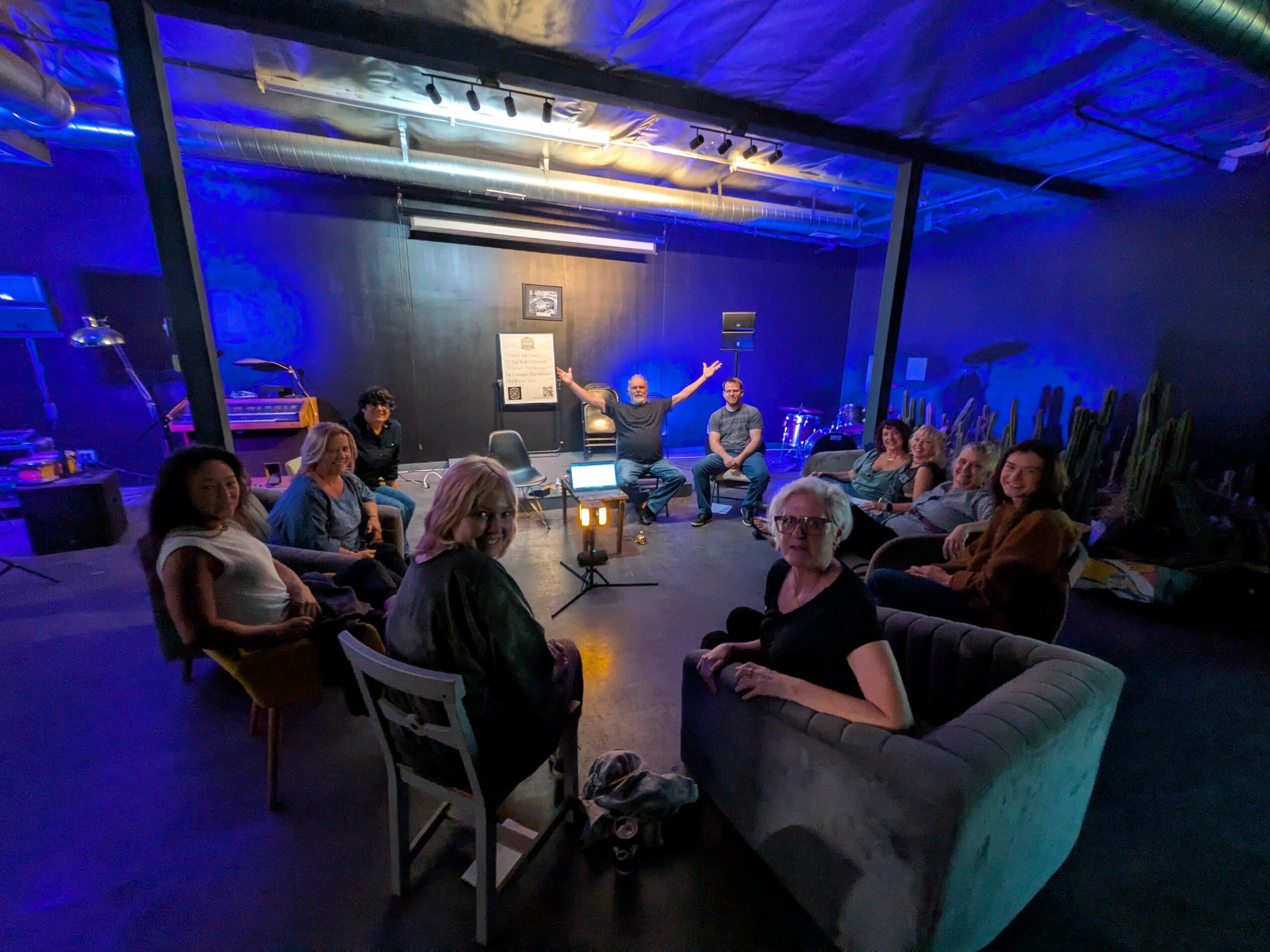 People seated in a circle in a dimly lit room with blue lighting, some with arms raised, engaging in a group activity.