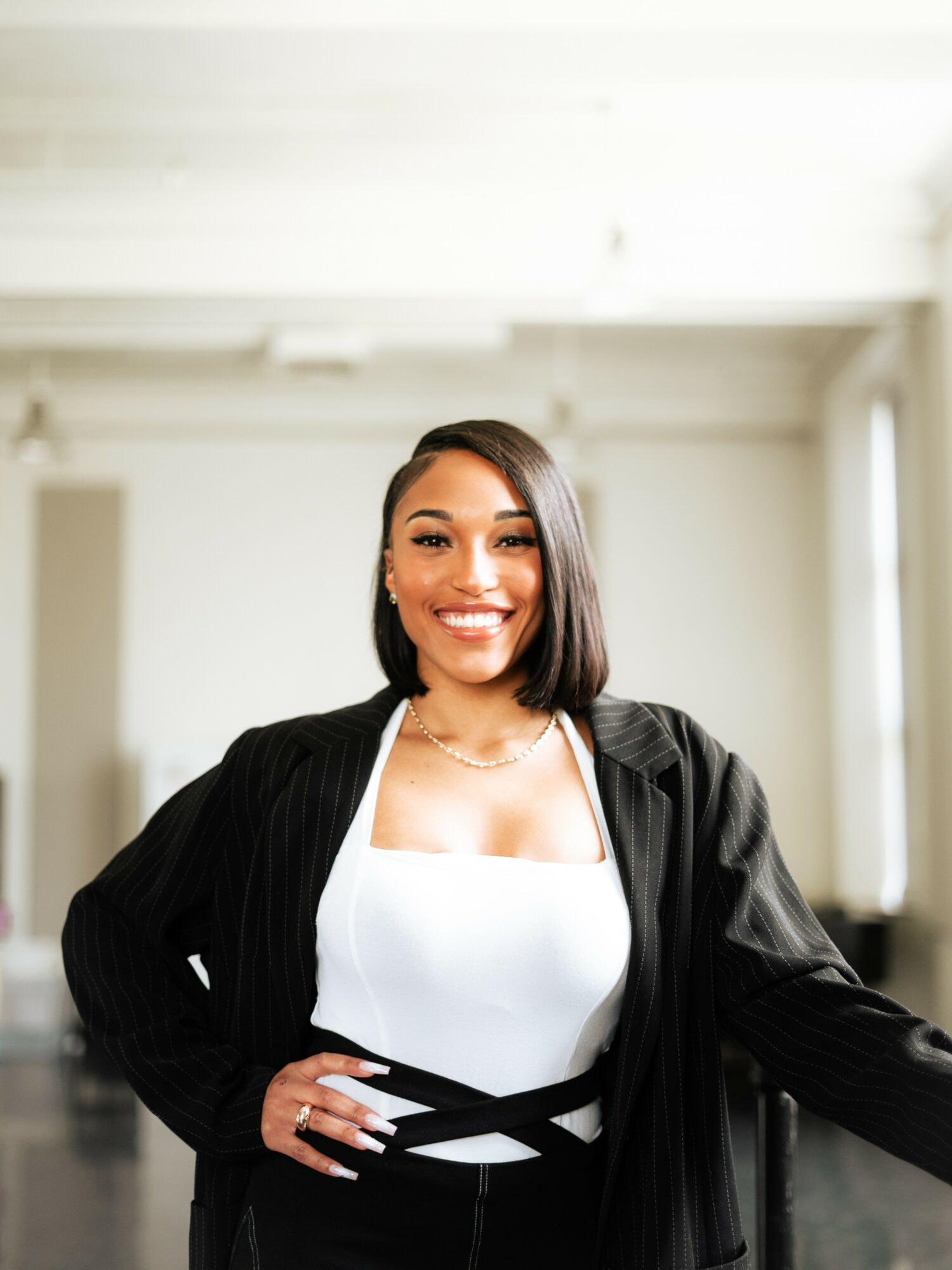 Smiling woman with shoulder-length hair, wearing a white top and black jacket, standing indoors with bright background.