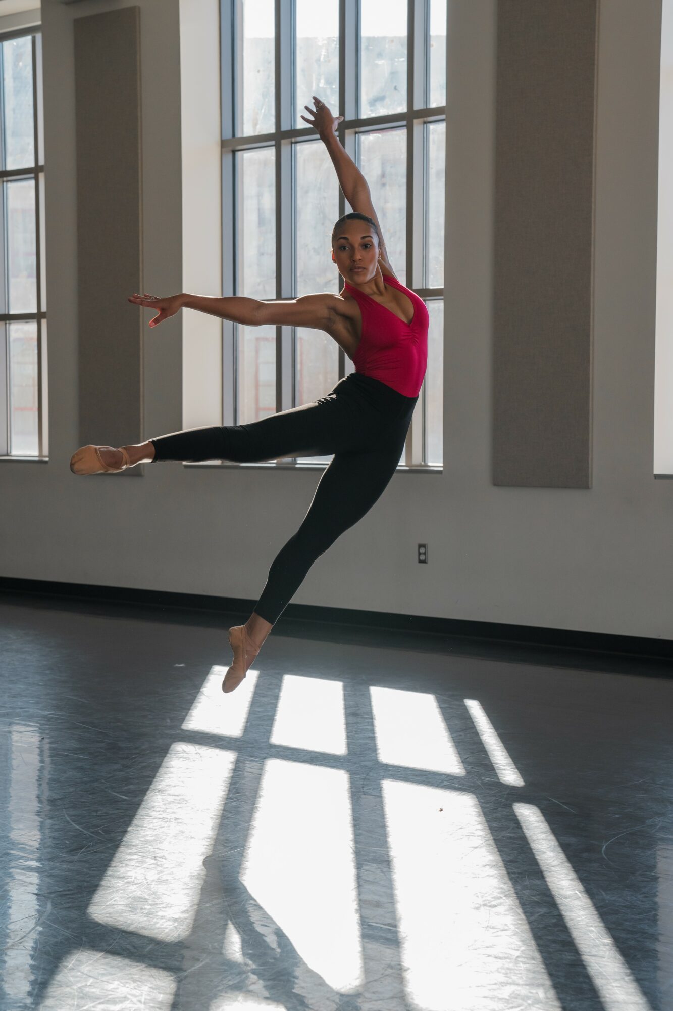 Ballet dancer in pink top and black pants jumps with arms and legs extended inside a studio with large windows.