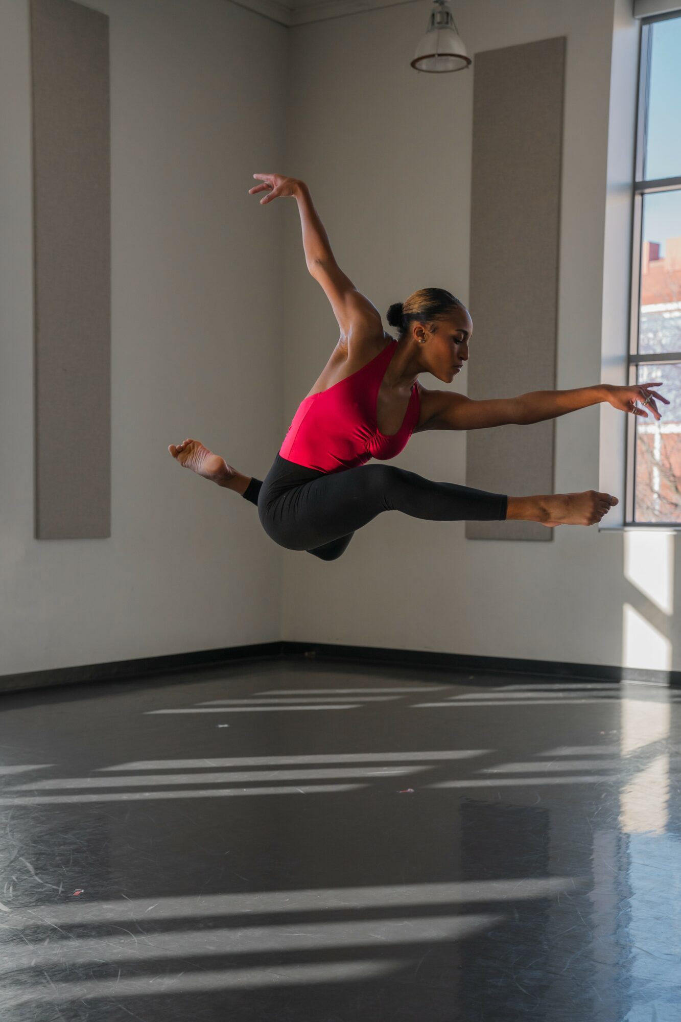 A woman in a pink top and black pants jumps in a room with large windows, sunlight casting shadows on the floor.