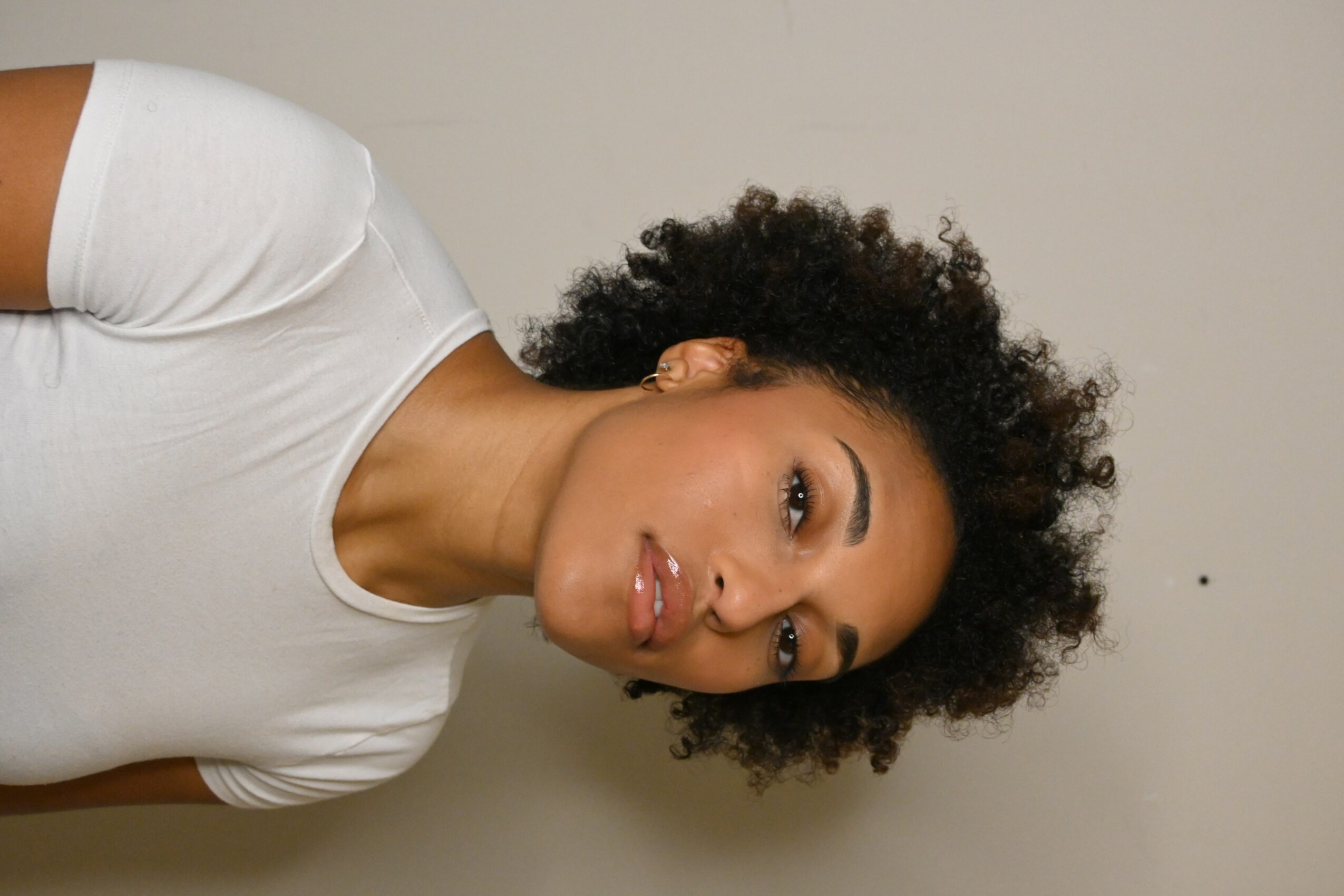 Woman with curly hair wearing a white shirt, lying sideways against a plain wall.