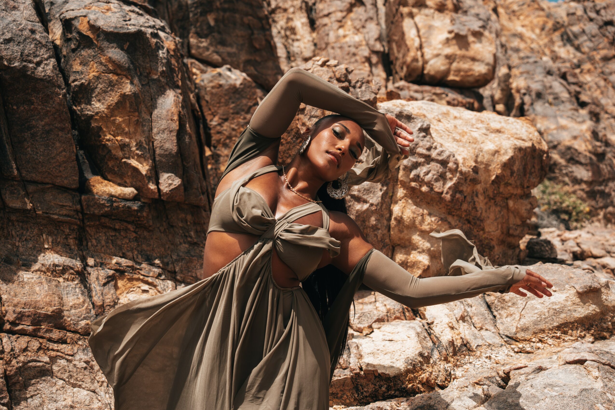 Woman in flowing dress poses against rocky background with arms raised above head, eyes closed, outdoors.