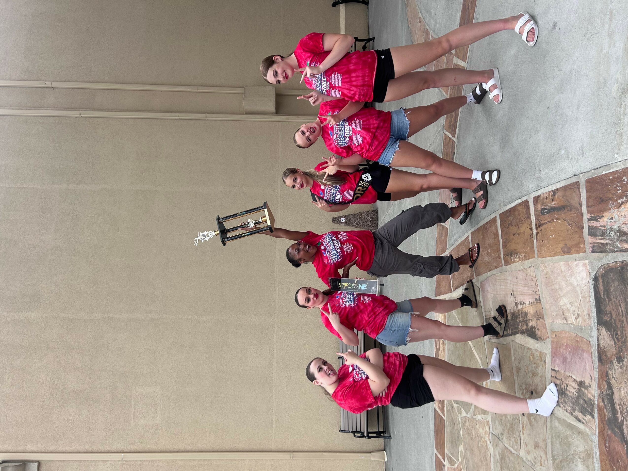 Group of six people in red shirts posing with a trophy, standing indoors against a plain wall.