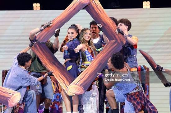 Group of young people holding large wooden frame, smiling and posing on stage with bright background.