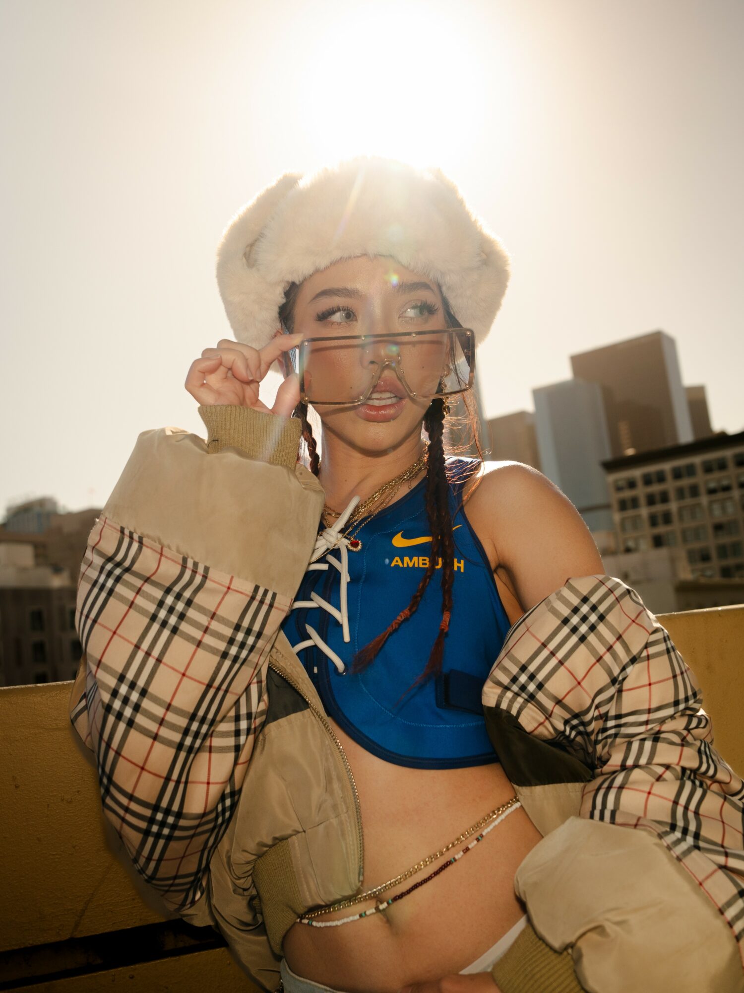 Young woman with glasses and a hat, wearing a blue top and beige jacket, standing outdoors with city buildings behind her.