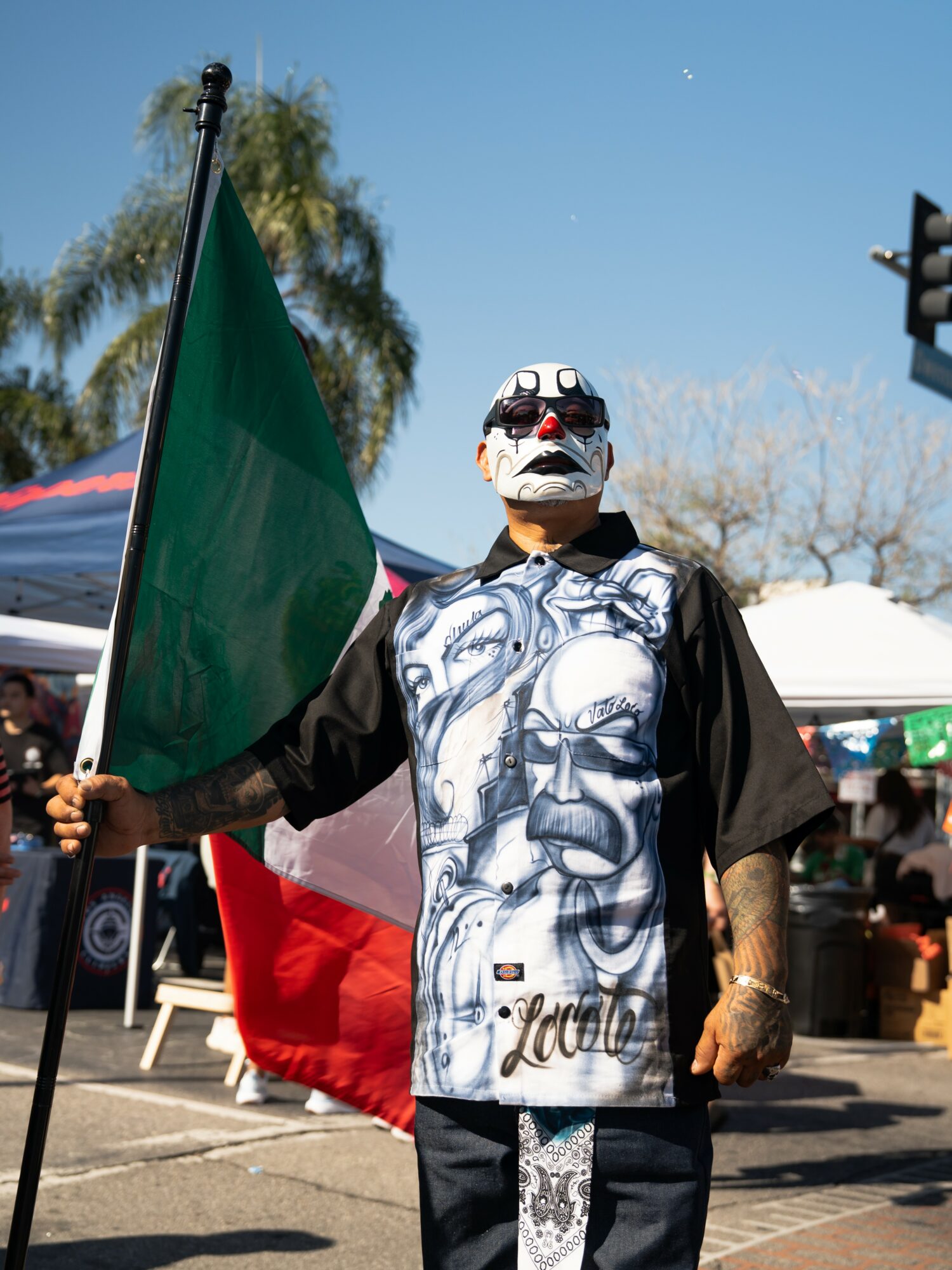 Person holding a flag, wearing a mask and a shirt with a face design, outdoors with tents and trees in background.