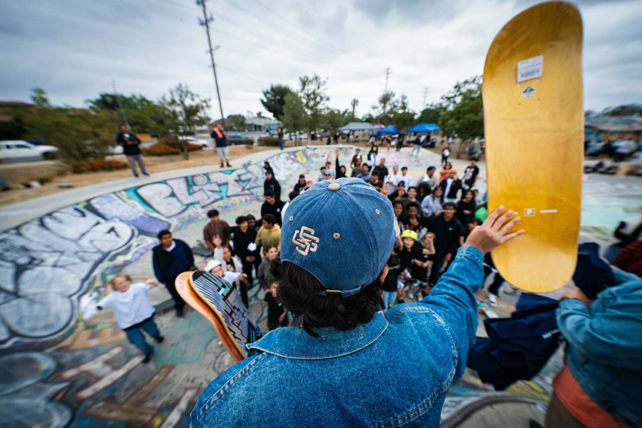 Crowd gathered at skate park with a person holding a skateboard, graffiti on the ramp, trees, and sky in background.