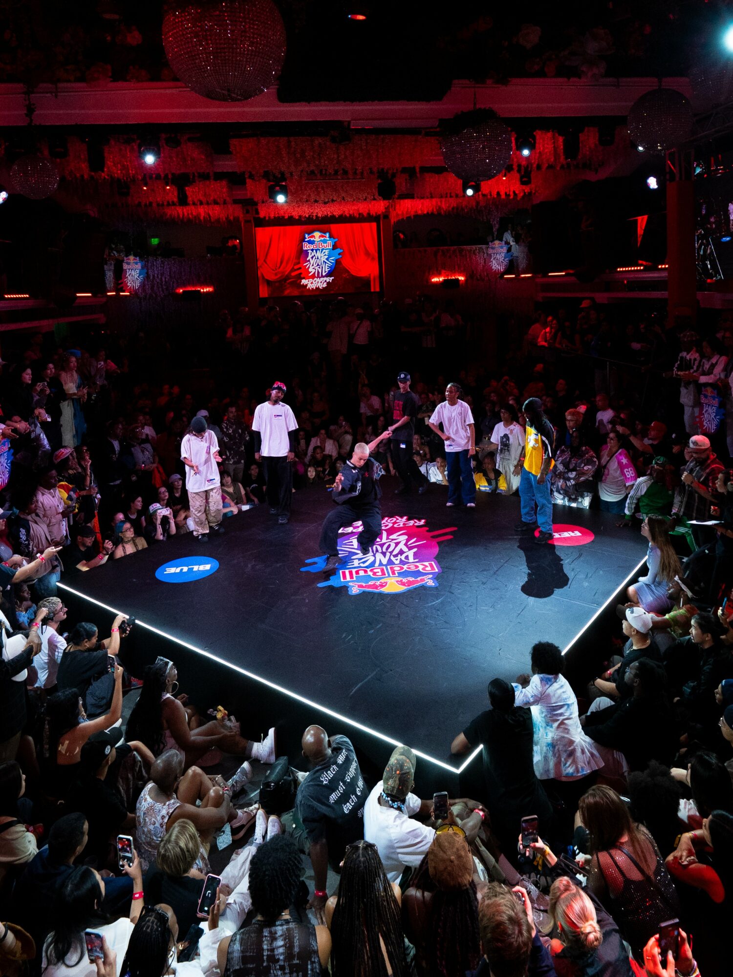 Dance battle on stage with performers and audience in a dark, red-lit venue, with a large screen above showing the scene.