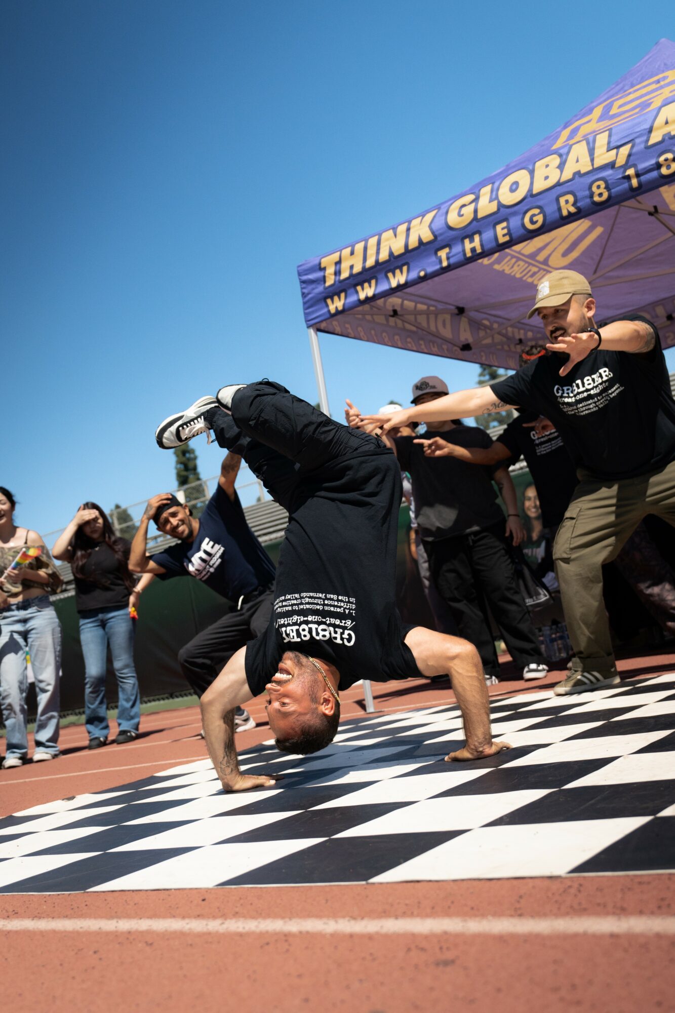 Person performing a headstand on a checkered floor at an outdoor event, with a crowd and a tent in the background.