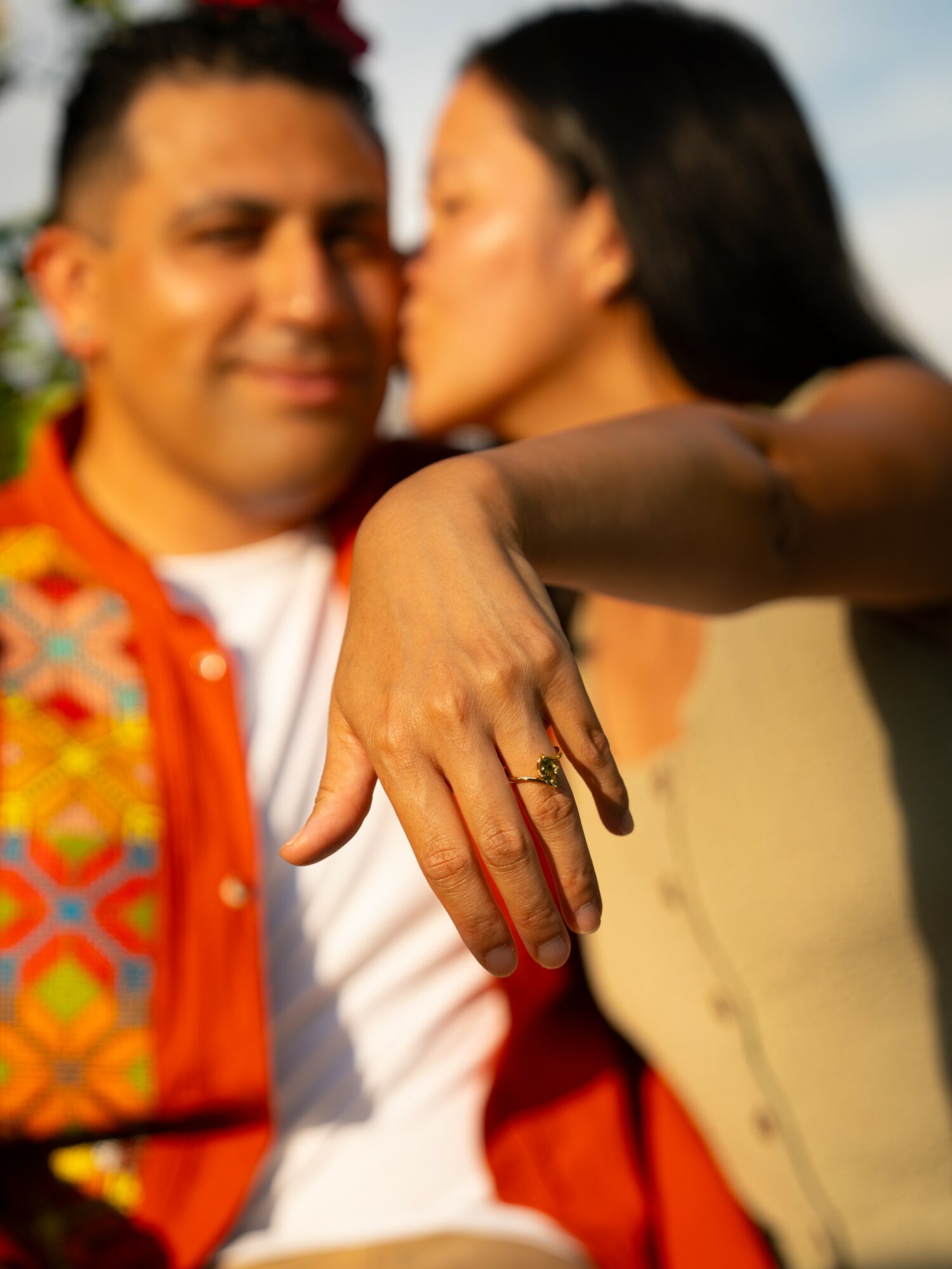 A woman shows her hand with a ring, with a man and woman in the background, outdoors, sunny day.