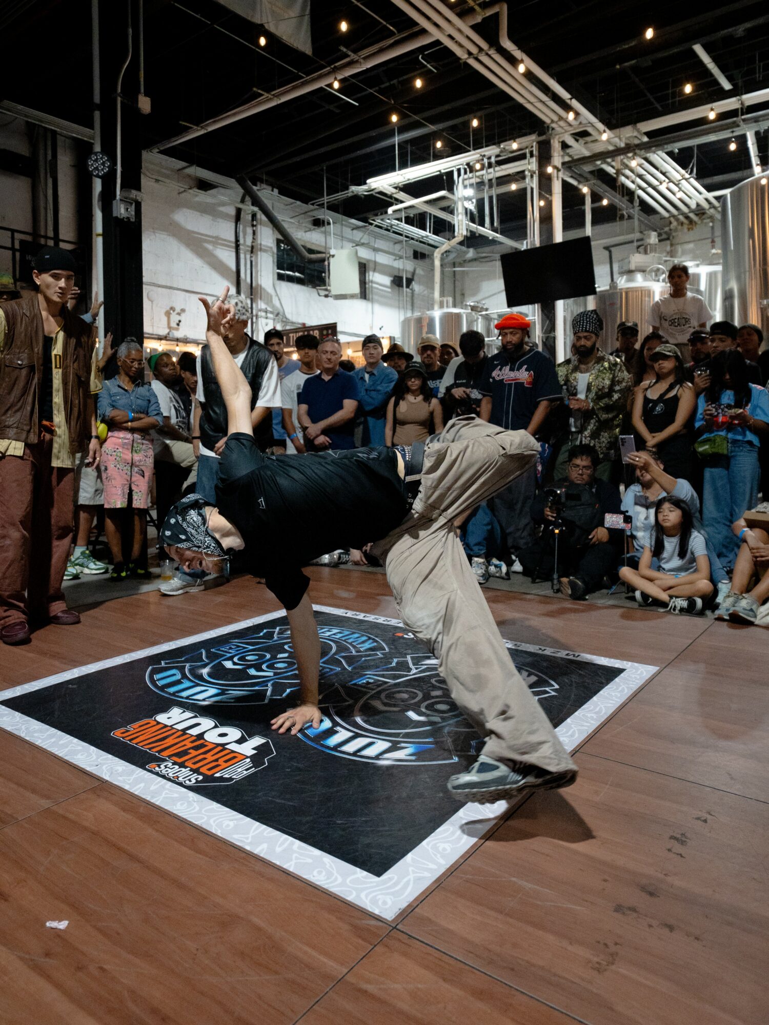 Person performing a breakdance move on a black mat with a crowd watching indoors.