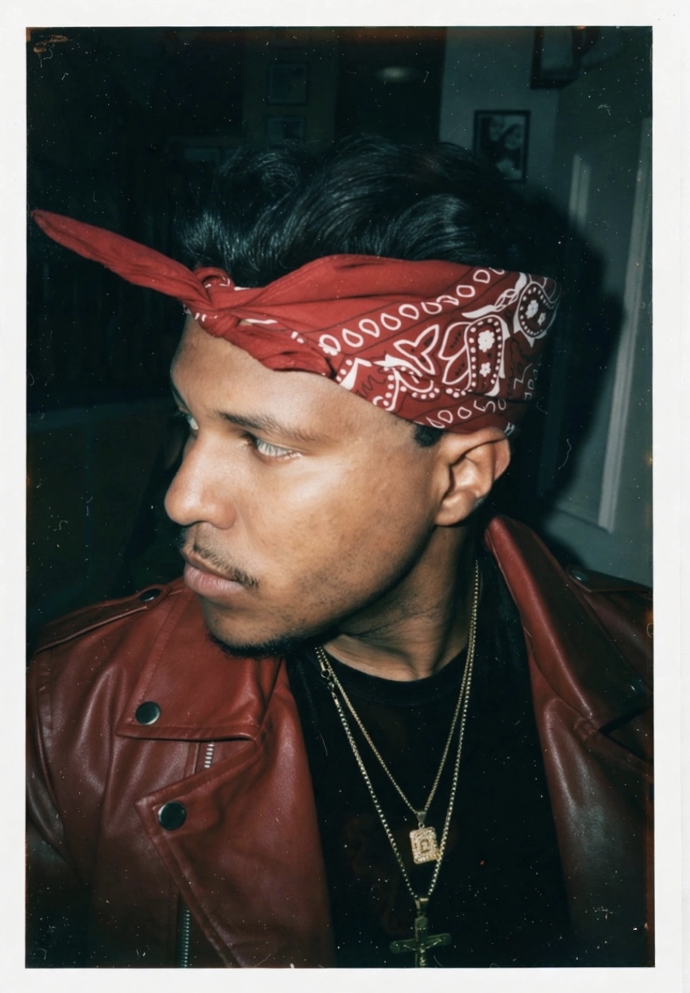 Young man with a red bandana, wearing a leather jacket and layered necklaces, looking to the side indoors.