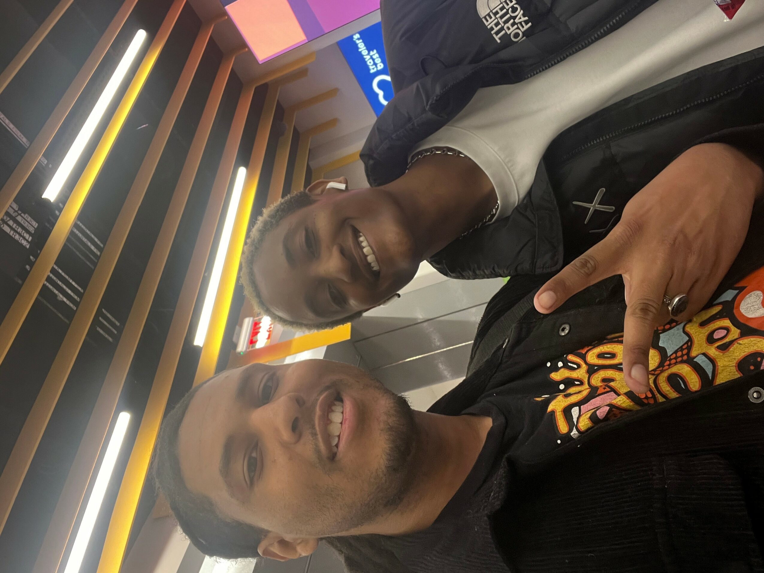 Two smiling men taking a selfie in an indoor setting with colorful lighting and wooden decor.